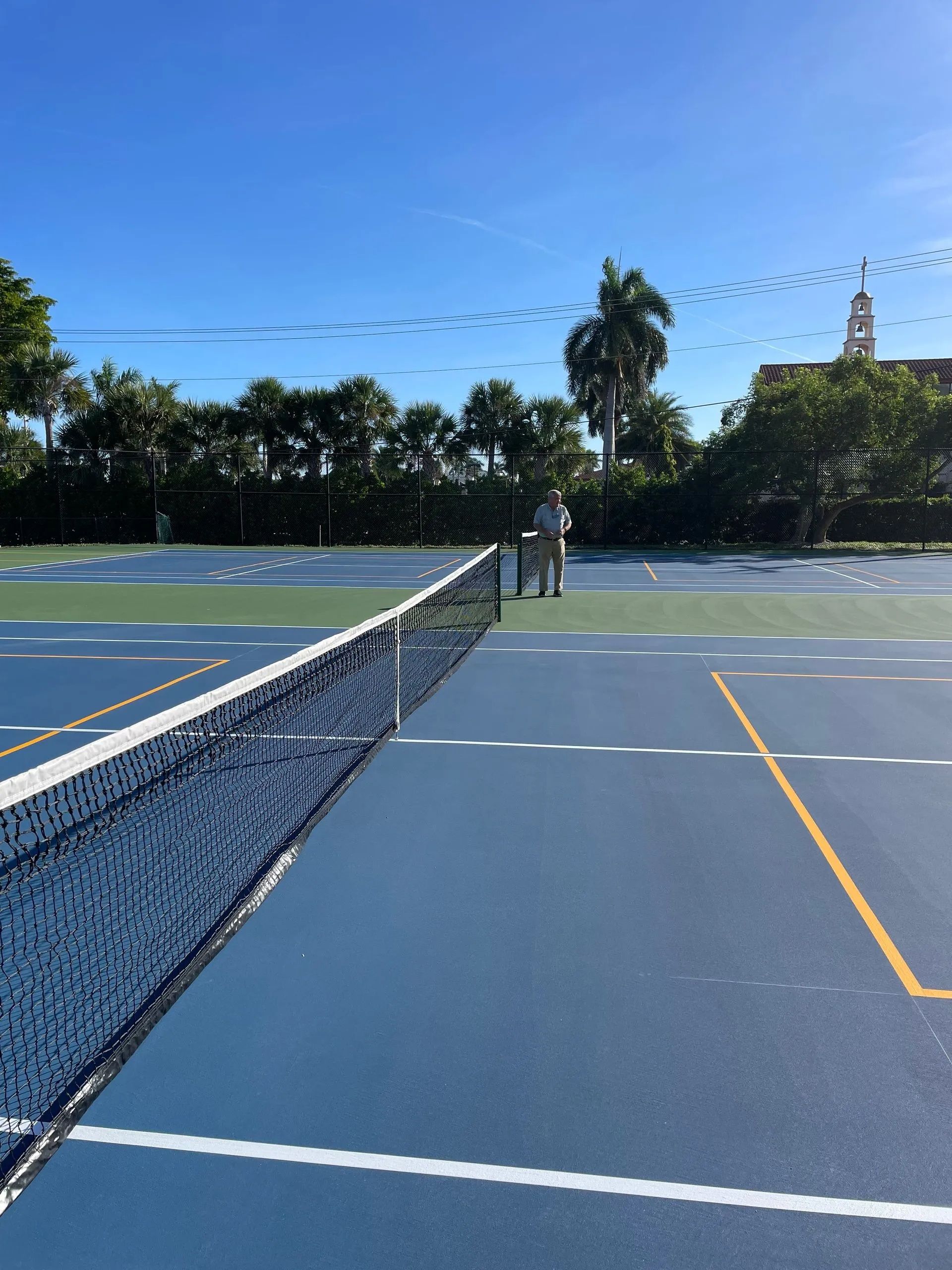 Blue outdoor tennis courts under a clear sky, with nets and trees in the background.
