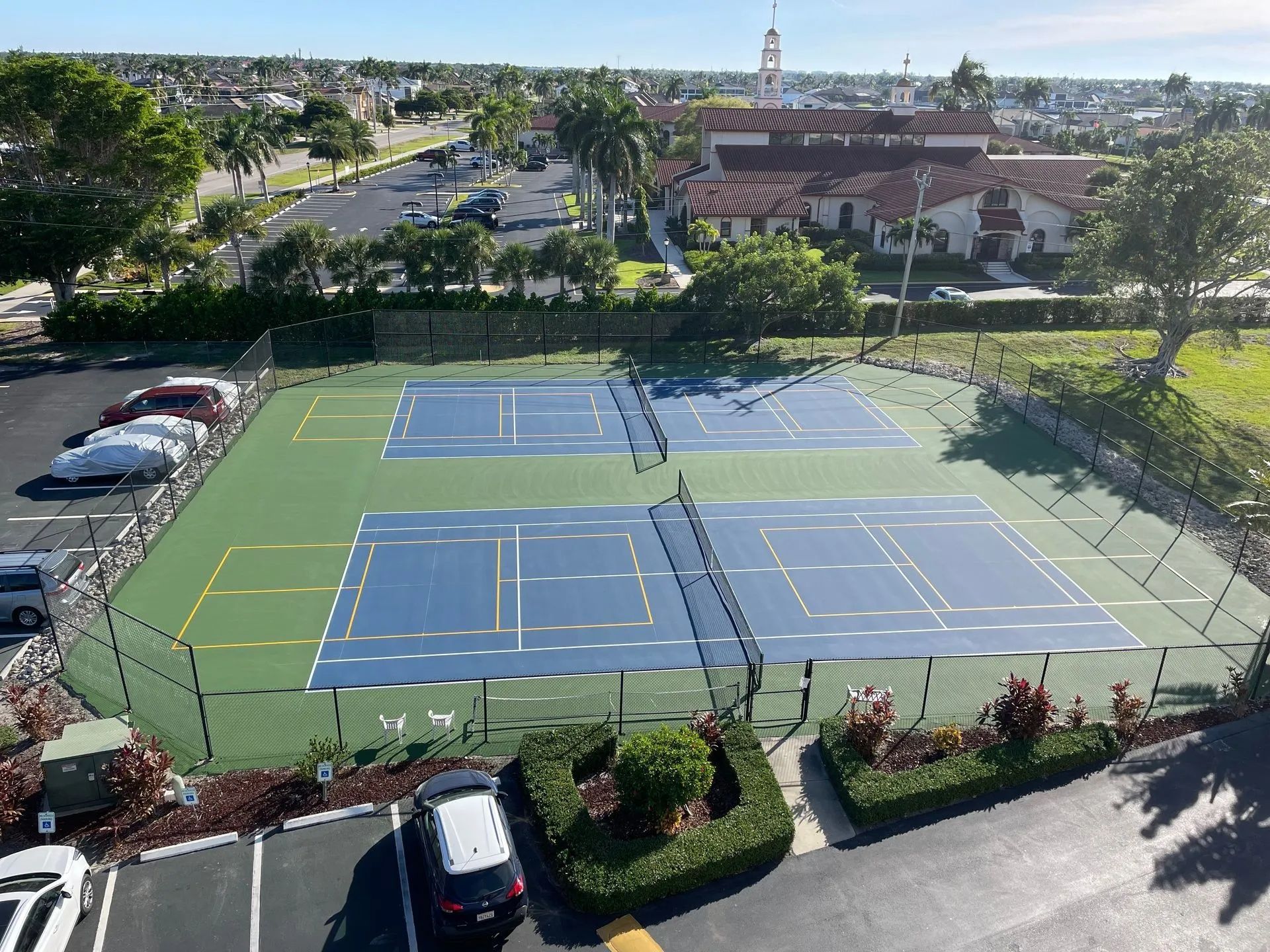 Aerial view of two blue tennis courts beside parking lots and landscaped greenery.