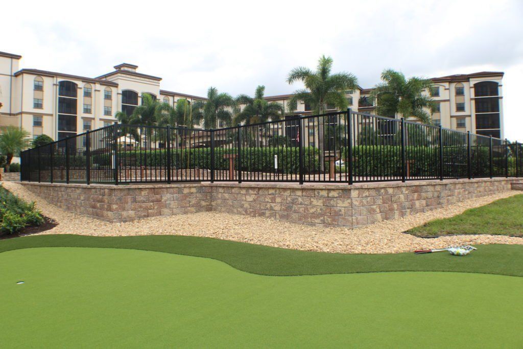 Resort-style apartment buildings behind a black fence and palm trees, beside a green pond