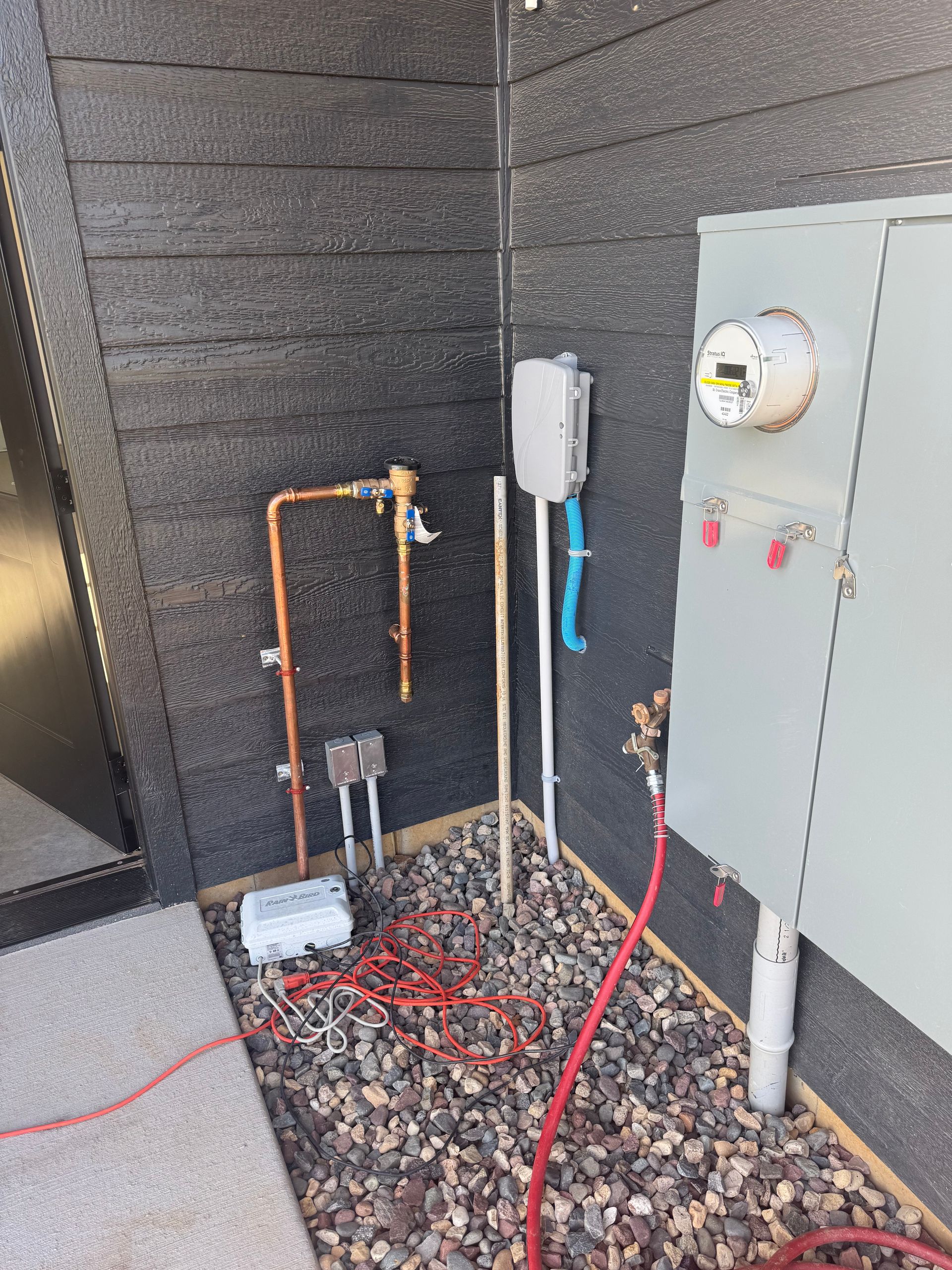 Outdoor utility equipment beside a building, with pipes, a meter, and electrical panel in a gravel bed.