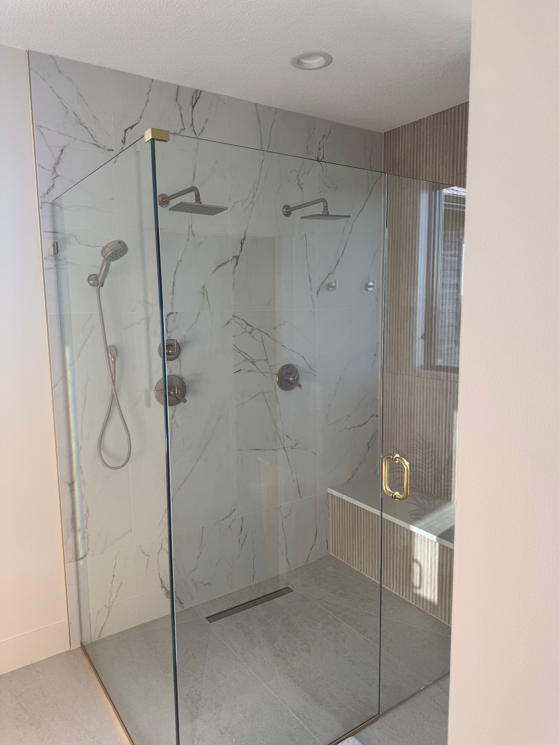 Modern marble-tiled walk-in shower with glass enclosure, rain showerhead, and a built-in bench by the window.