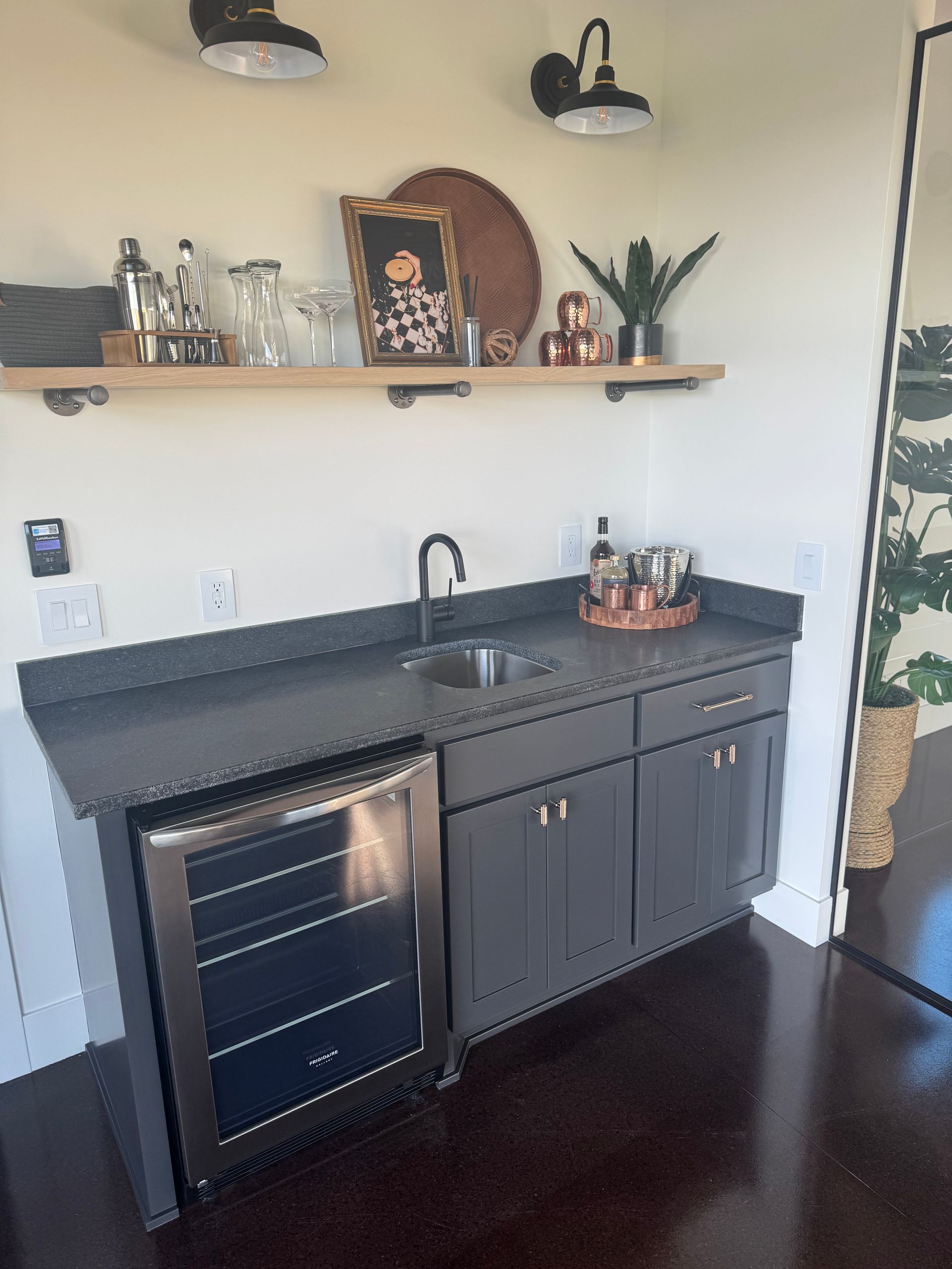 Small home bar with black countertop, mini fridge, sink, shelves, and decor on the wall.
