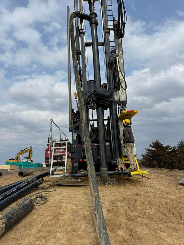 Worker operating a tall drilling rig at a construction site with hoses and equipment on muddy ground.