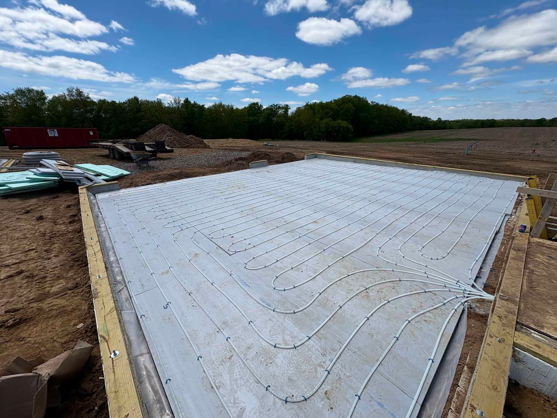 White concrete foundation slab at a construction site under a blue sky, with dirt and trees around it