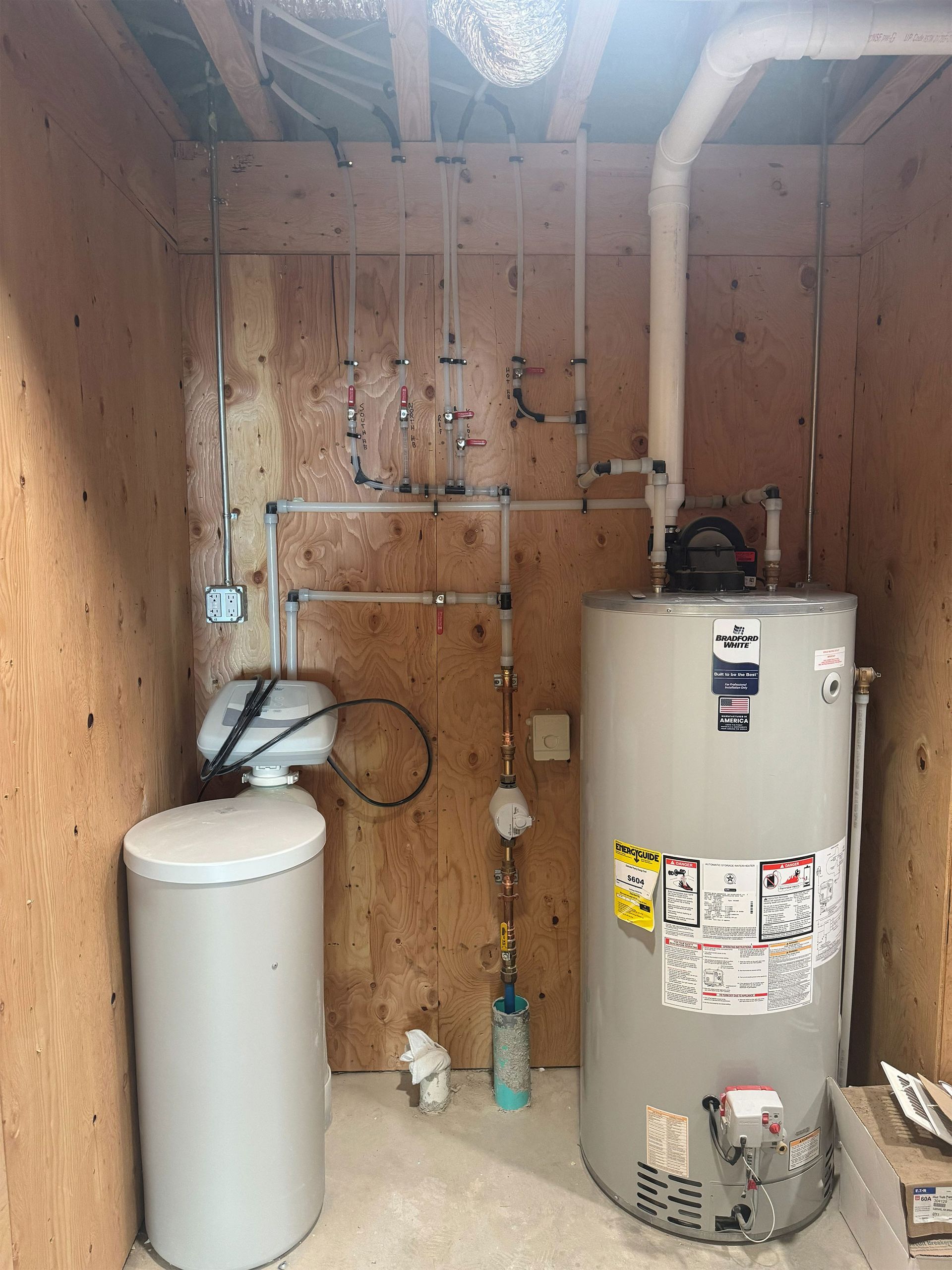Basement utility room with water heater, pressure tank, and exposed pipes against plywood walls
