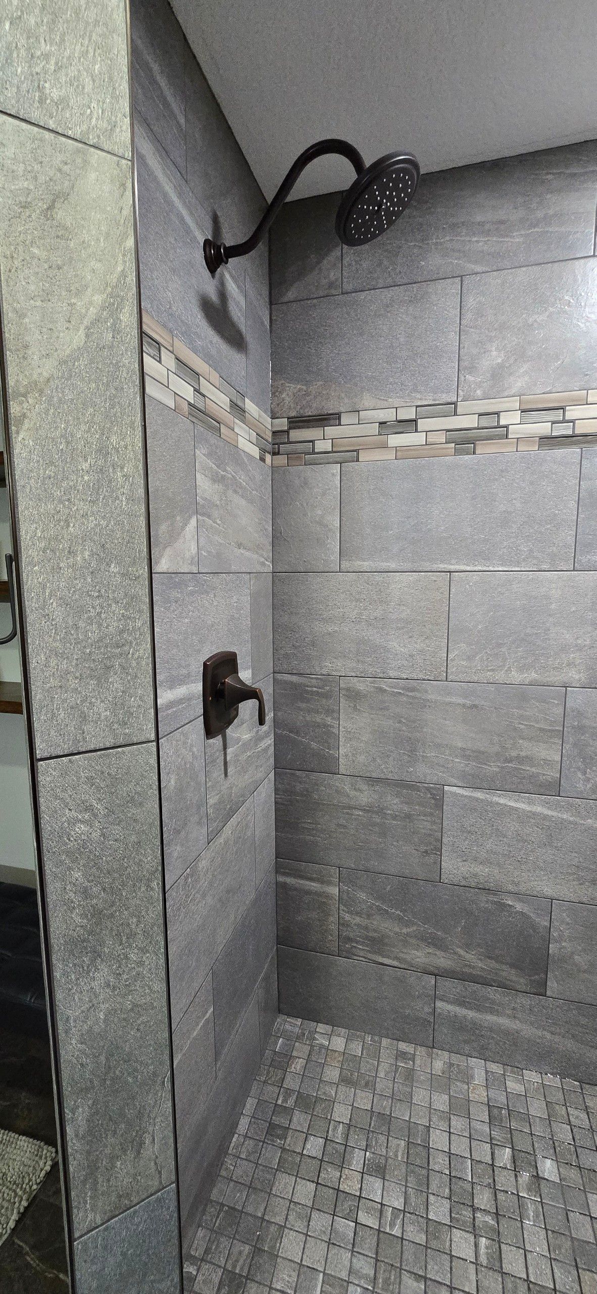 Gray tiled shower with a black showerhead and soap dispenser on the wall