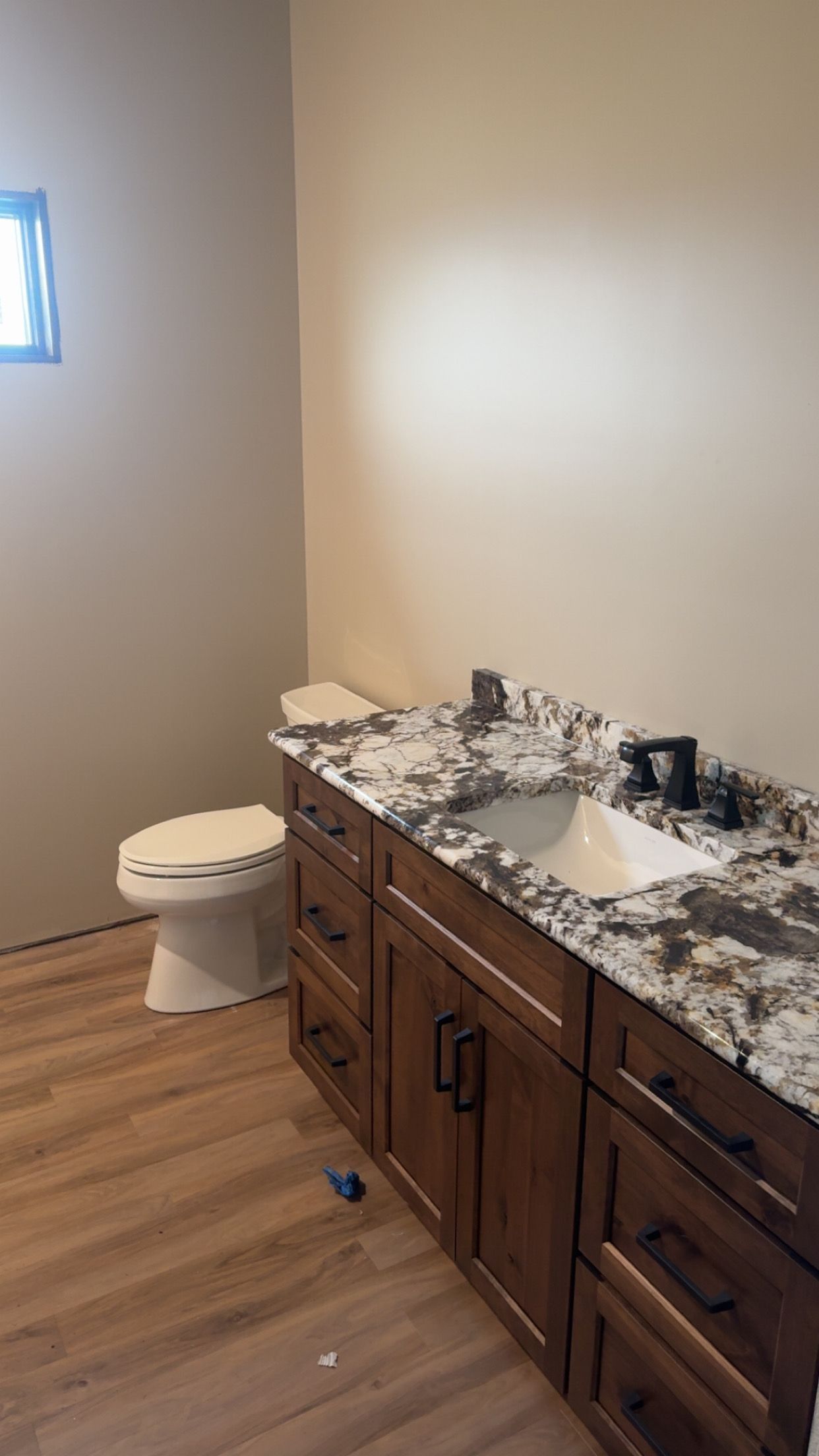 Bathroom with wood vanity, speckled countertop, white sink, toilet, and small window.