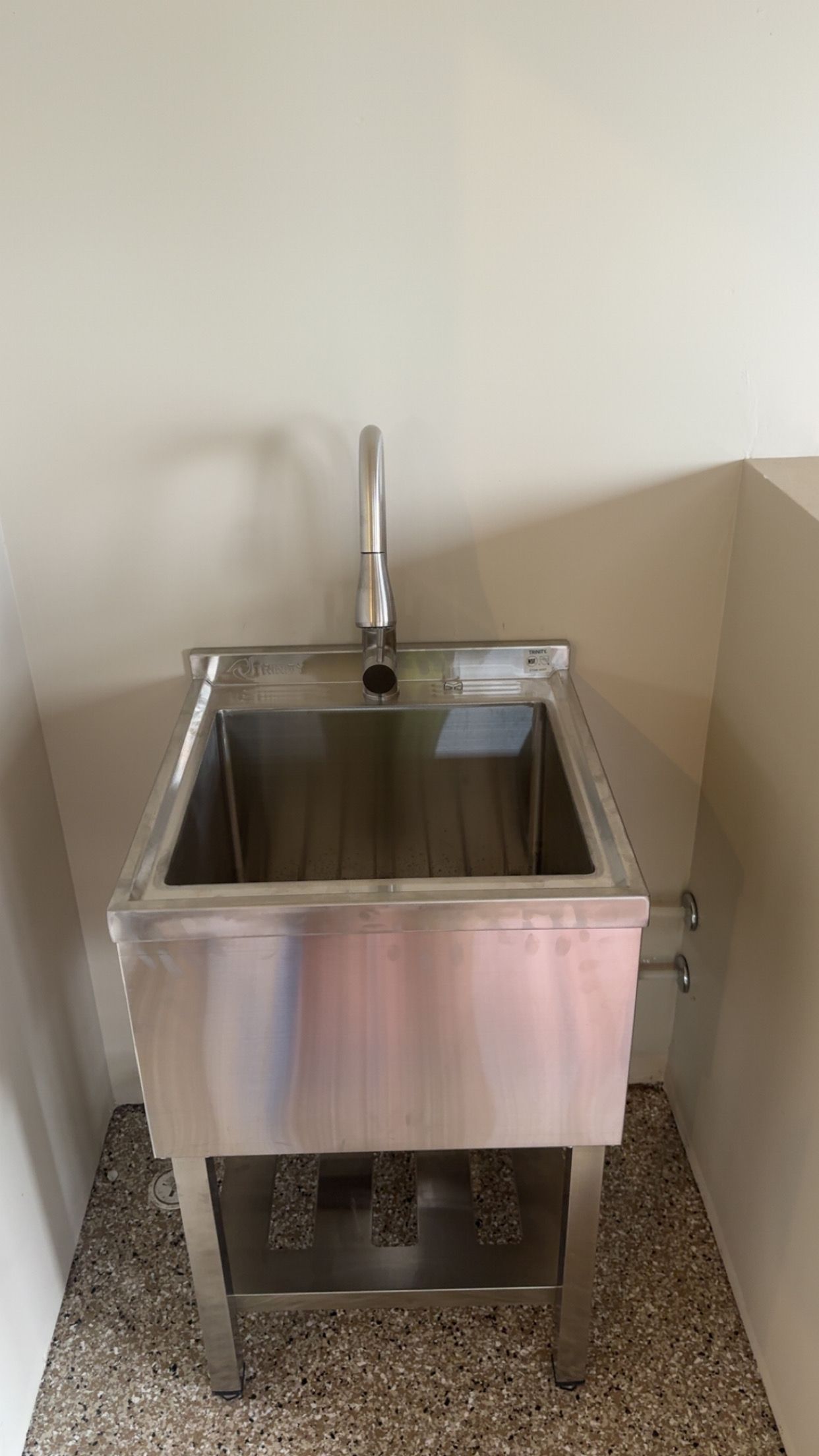 Stainless steel sink with faucet in a corner, mounted on metal legs over a speckled floor.