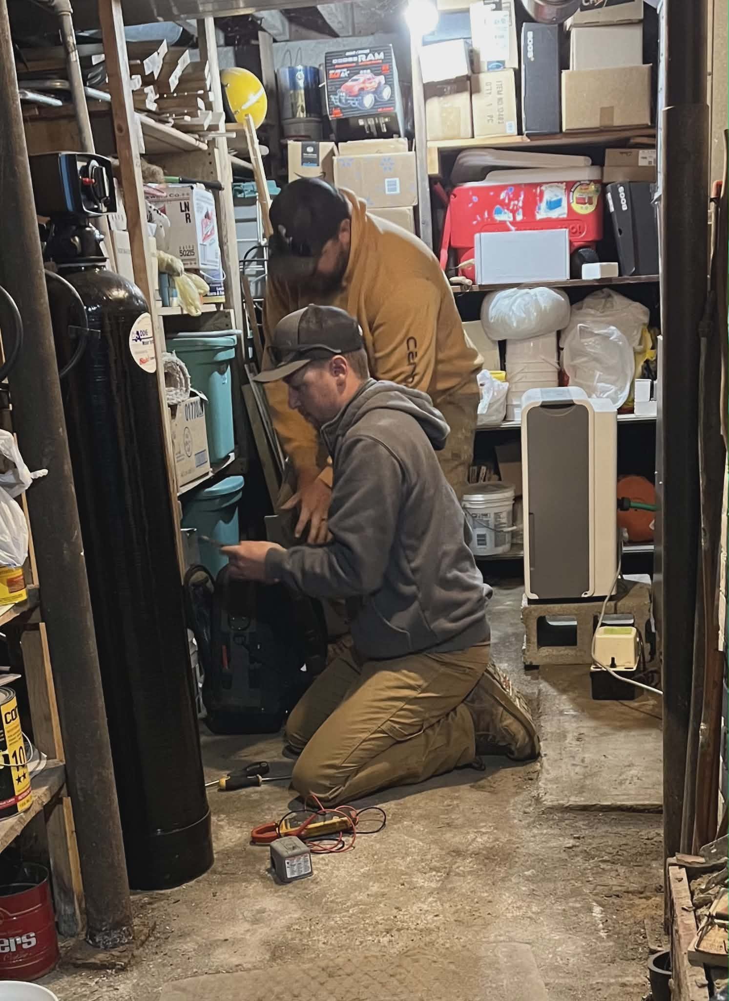 Two people kneel in a cluttered workshop, working on a vertical machine amid shelves and boxes.