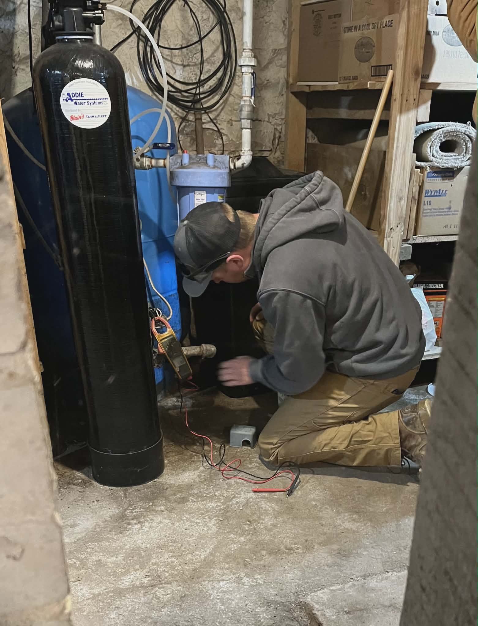 Technician kneels beside a blue tank, working in a cluttered basement utility room.