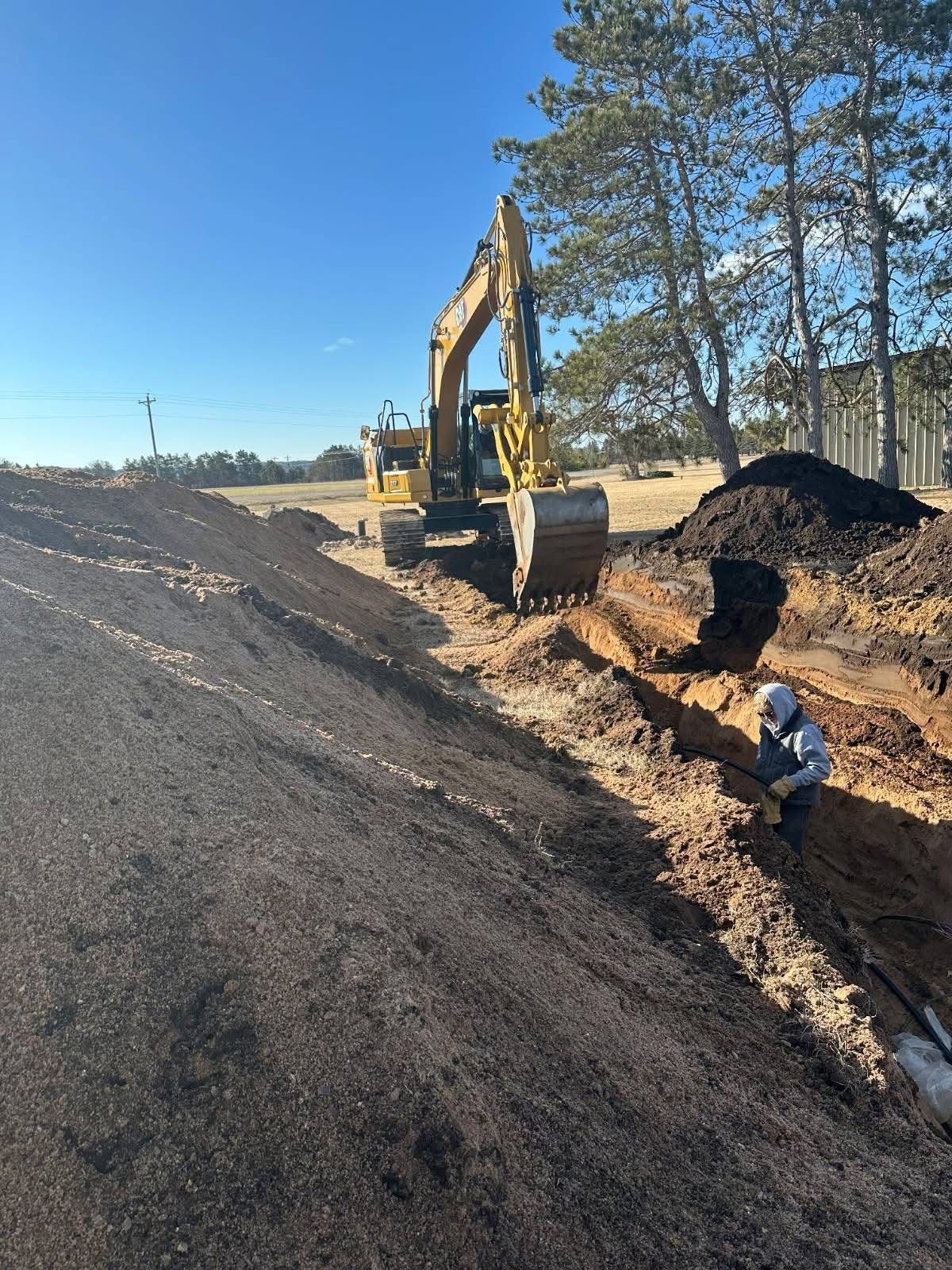 Excavator digging a trench beside a dirt road under a clear blue sky, with trees in the background