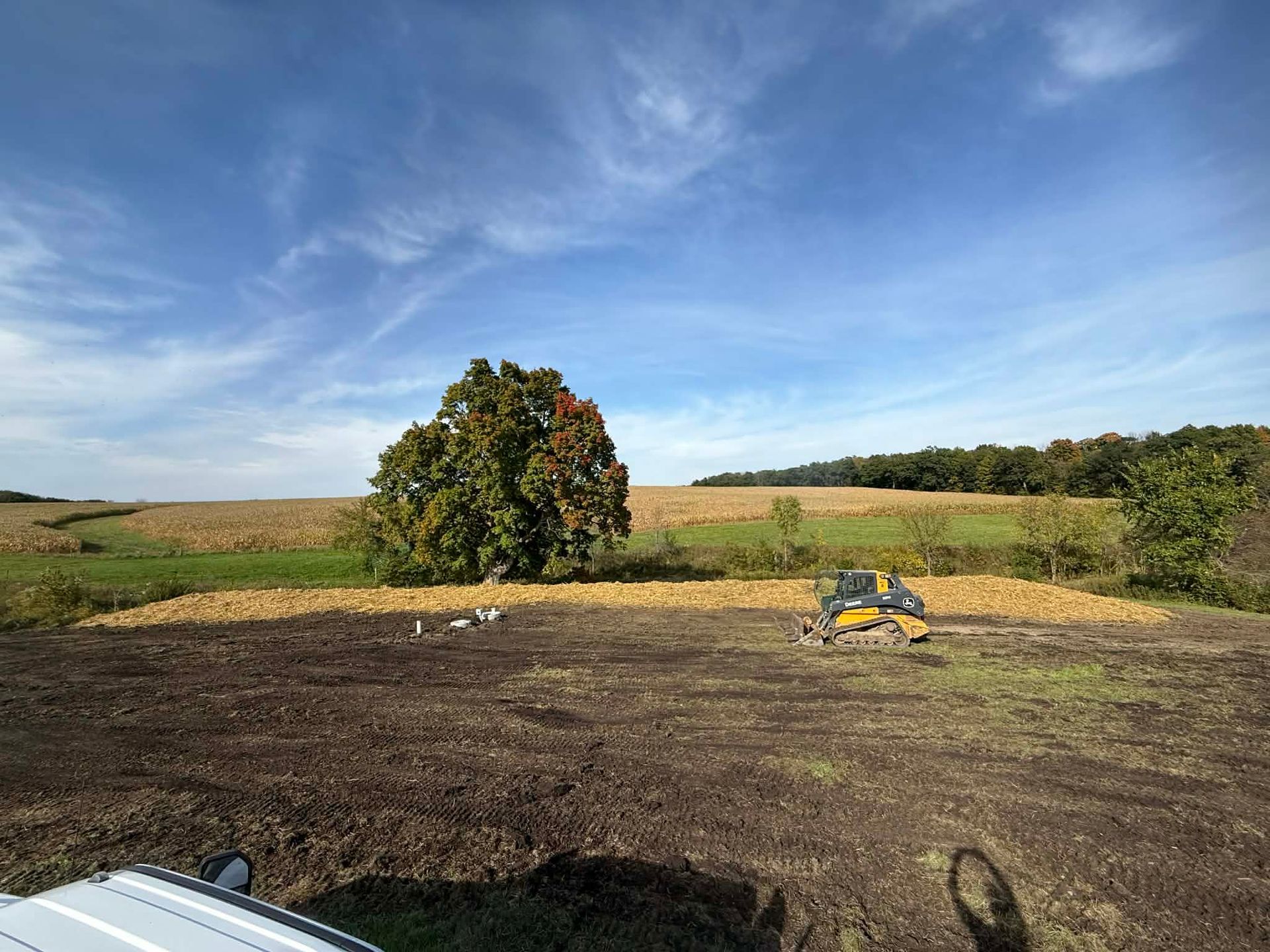 Open field with a large tree, dark soil, and a small yellow machine under a blue sky.