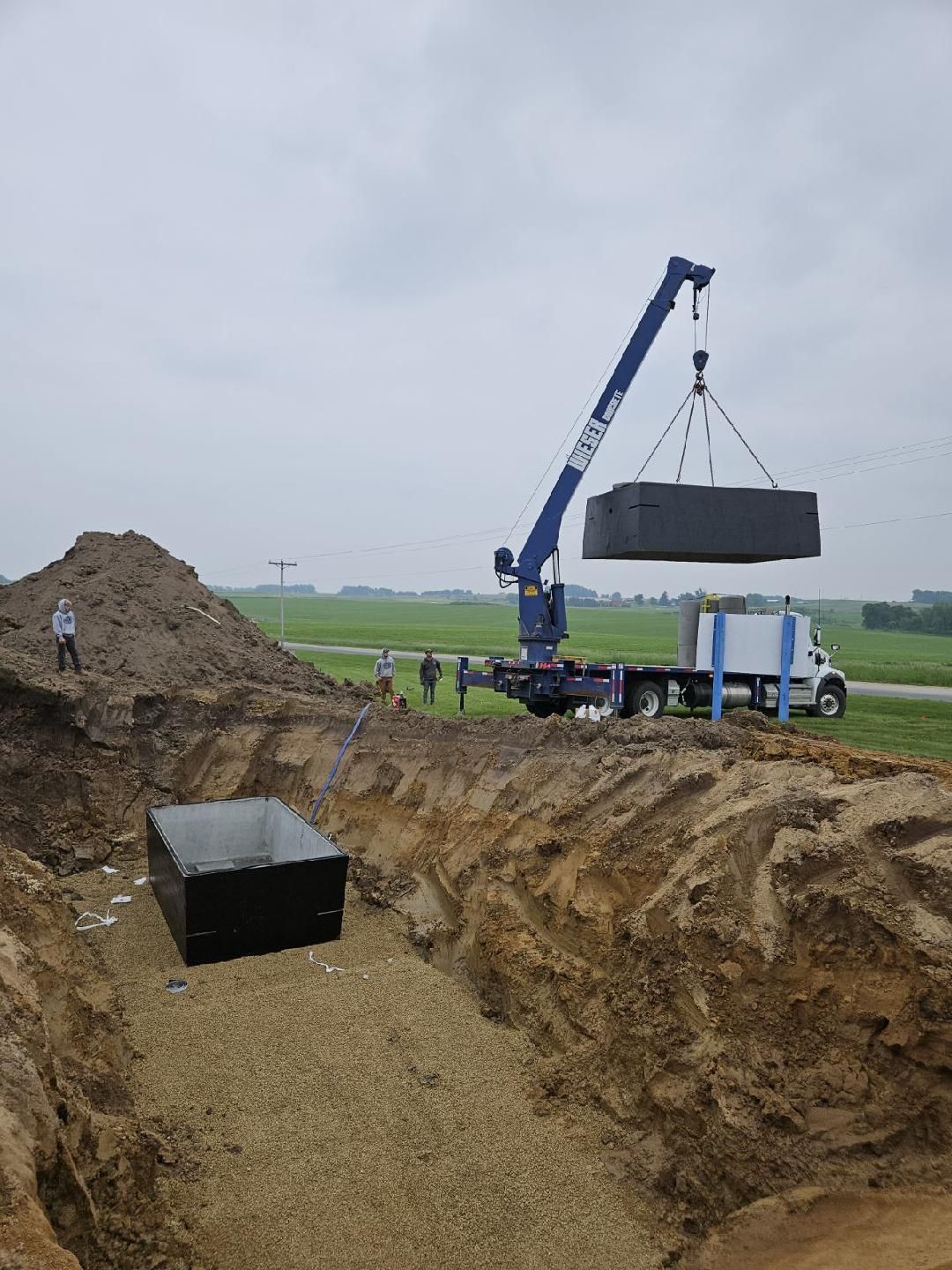 Excavator lifting a large concrete block into a trench at a construction site.