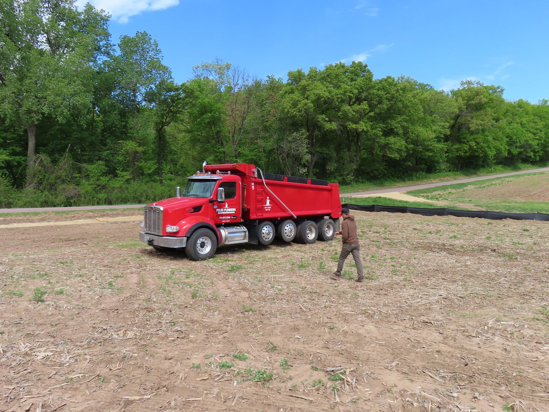 Red dump truck on a dirt field with a person walking nearby, trees and blue sky in the background