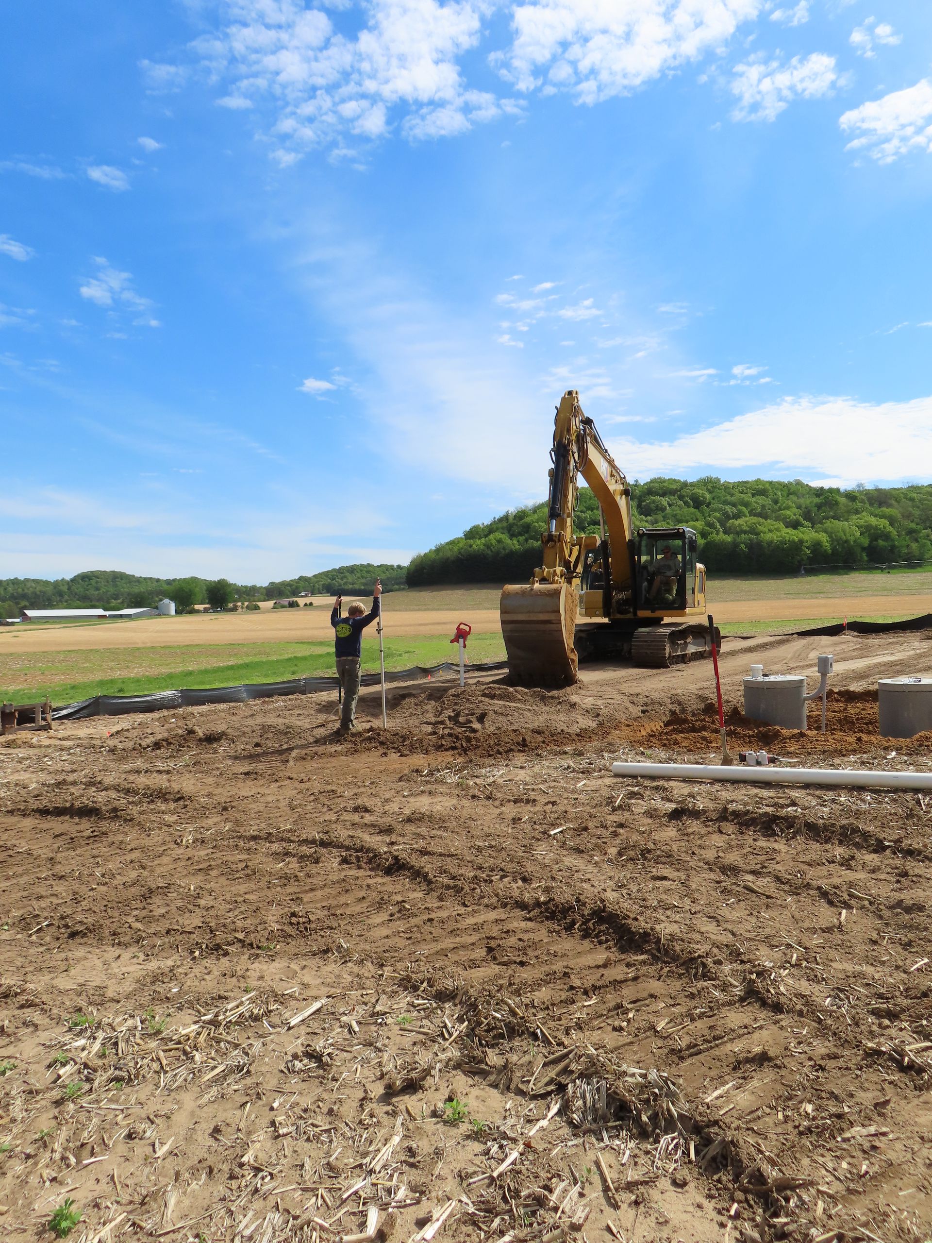 Excavator and worker at a dirt construction site under a bright blue sky