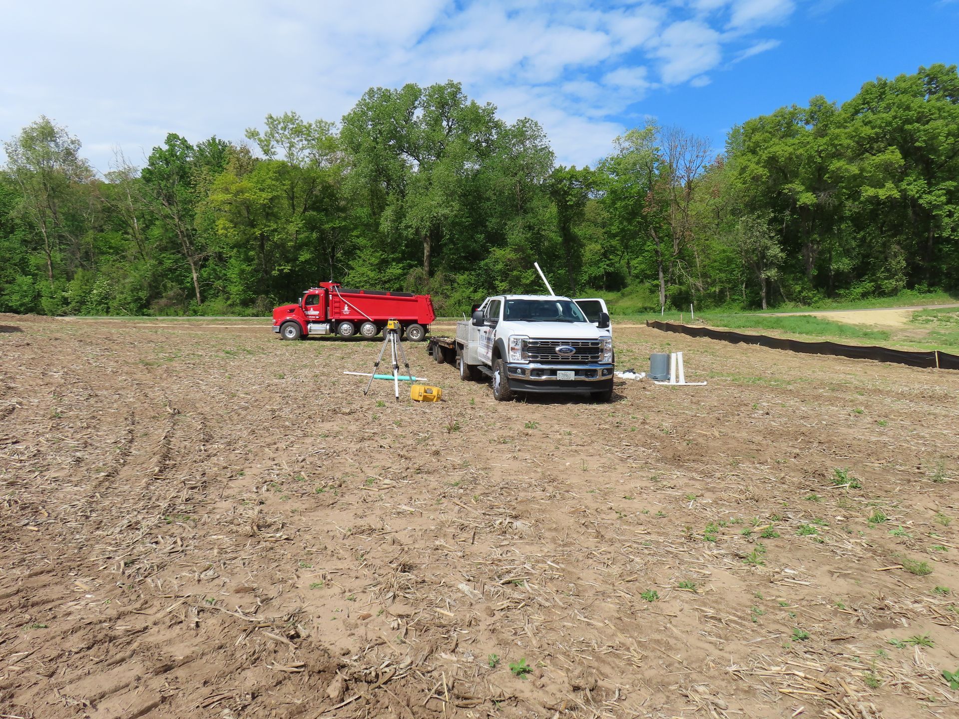 White pickup and red truck in a plowed field with markers, trees and blue sky in the background
