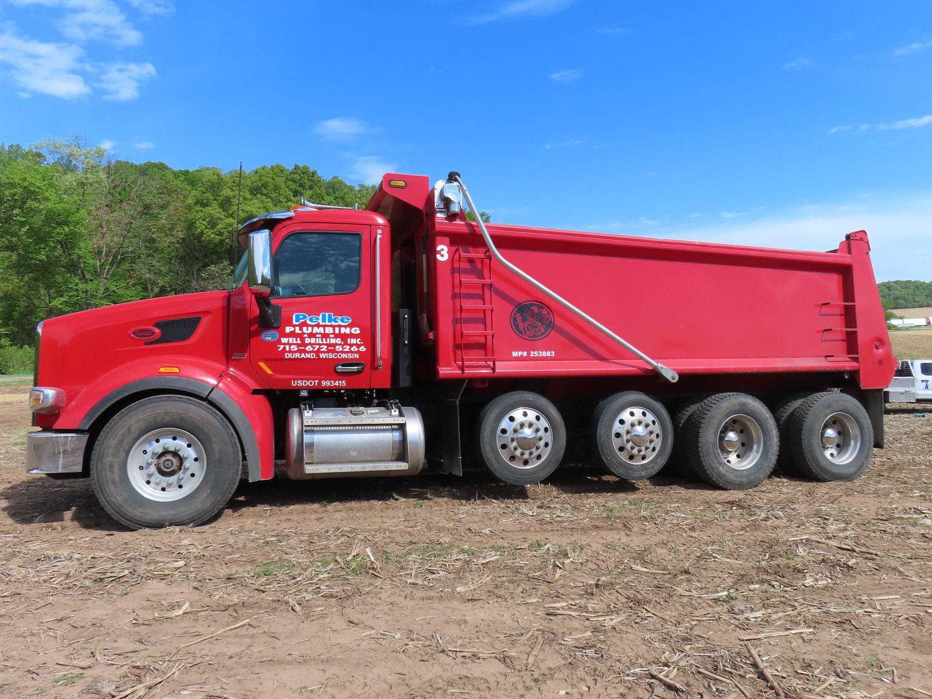 Red dump truck parked on dirt under a blue sky with trees in the background