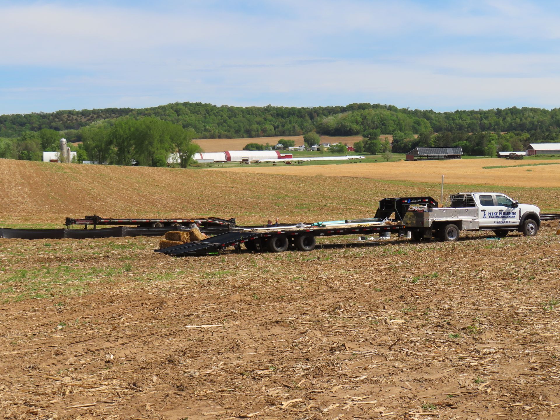White pickup truck towing a long flatbed trailer across a harvested field under a blue sky