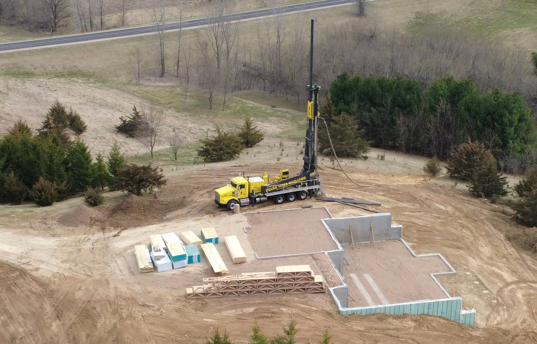 Yellow drill rig on a construction site with concrete foundations and materials laid out on bare ground.