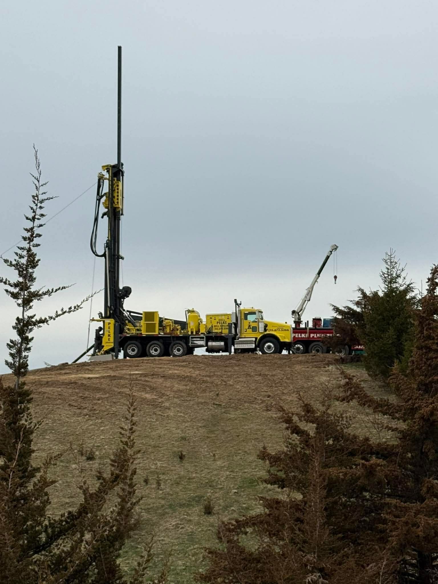 Yellow drilling rig and crane on a hill under a gray sky, with evergreen trees in the foreground