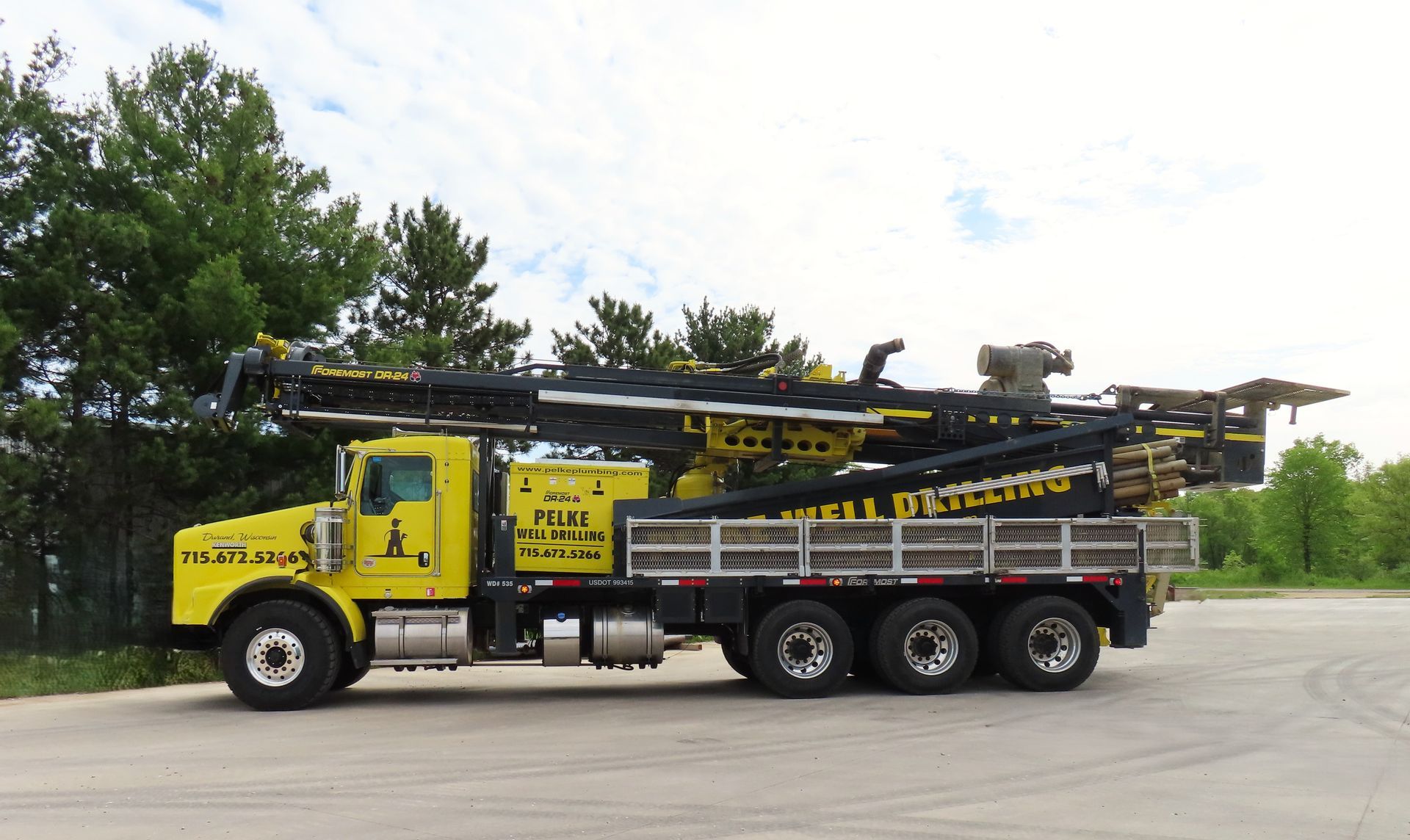 Yellow semi-truck carrying a black trailer with folded boom arms, parked outdoors on pavement.