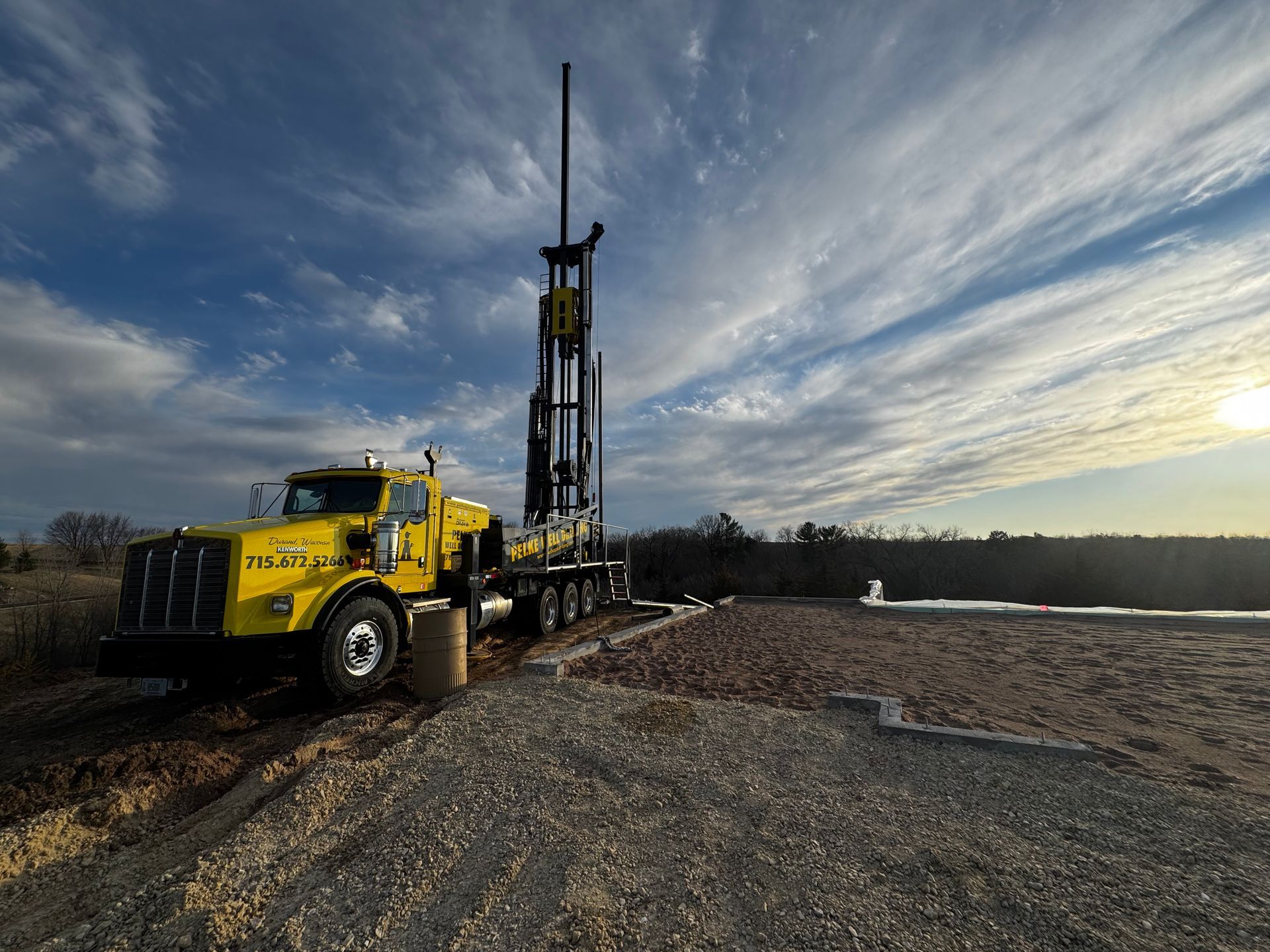 Yellow drilling rig truck on a gravel site at sunset, with a tall mast against a cloudy sky