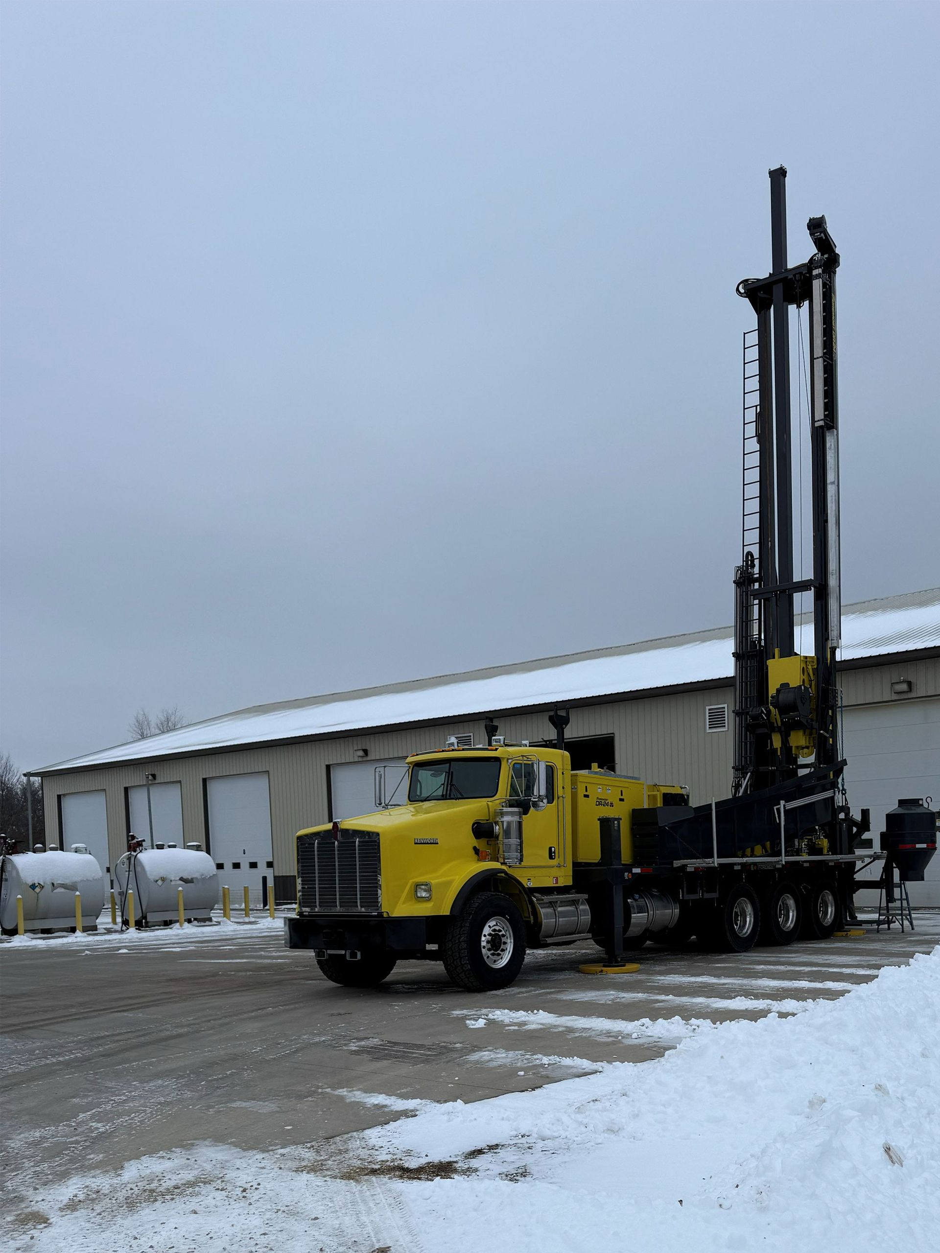 Yellow drilling truck beside a warehouse in a snowy industrial yard
