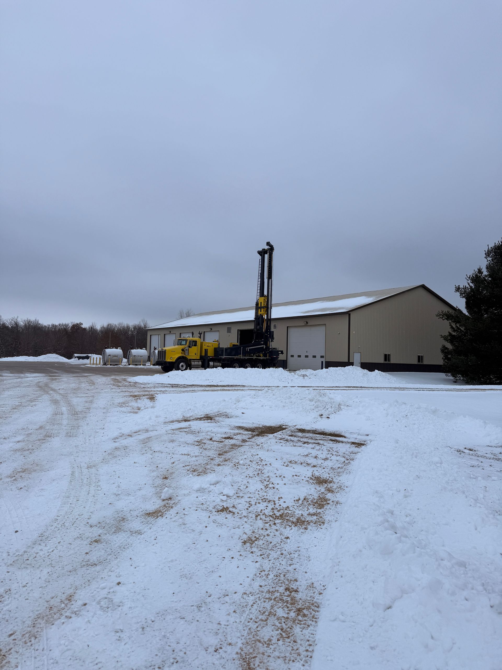 Snowy industrial yard with a yellow drilling rig beside a beige warehouse under an overcast sky