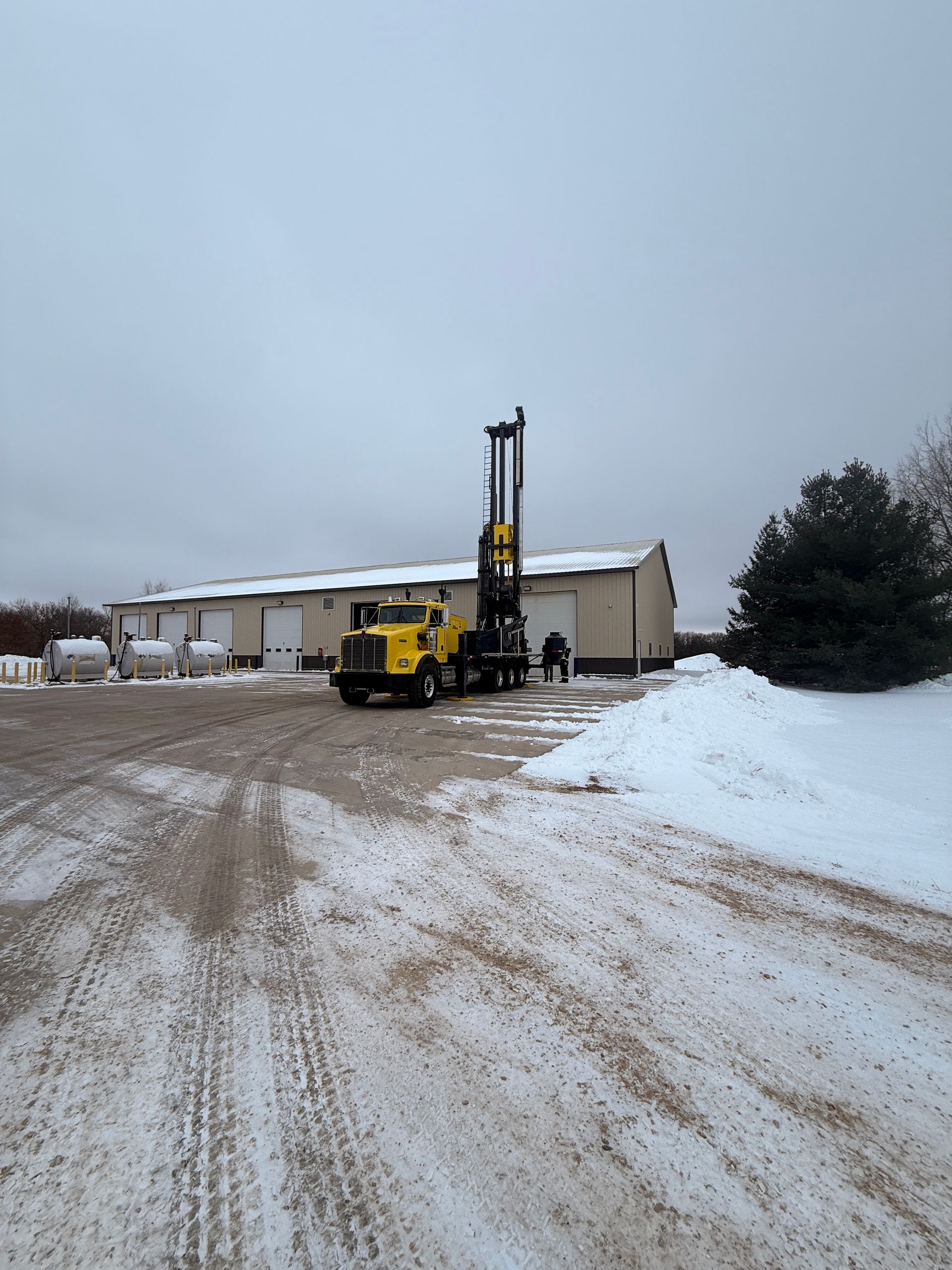 Yellow drilling rig on a snowy dirt lot beside a beige building under an overcast sky