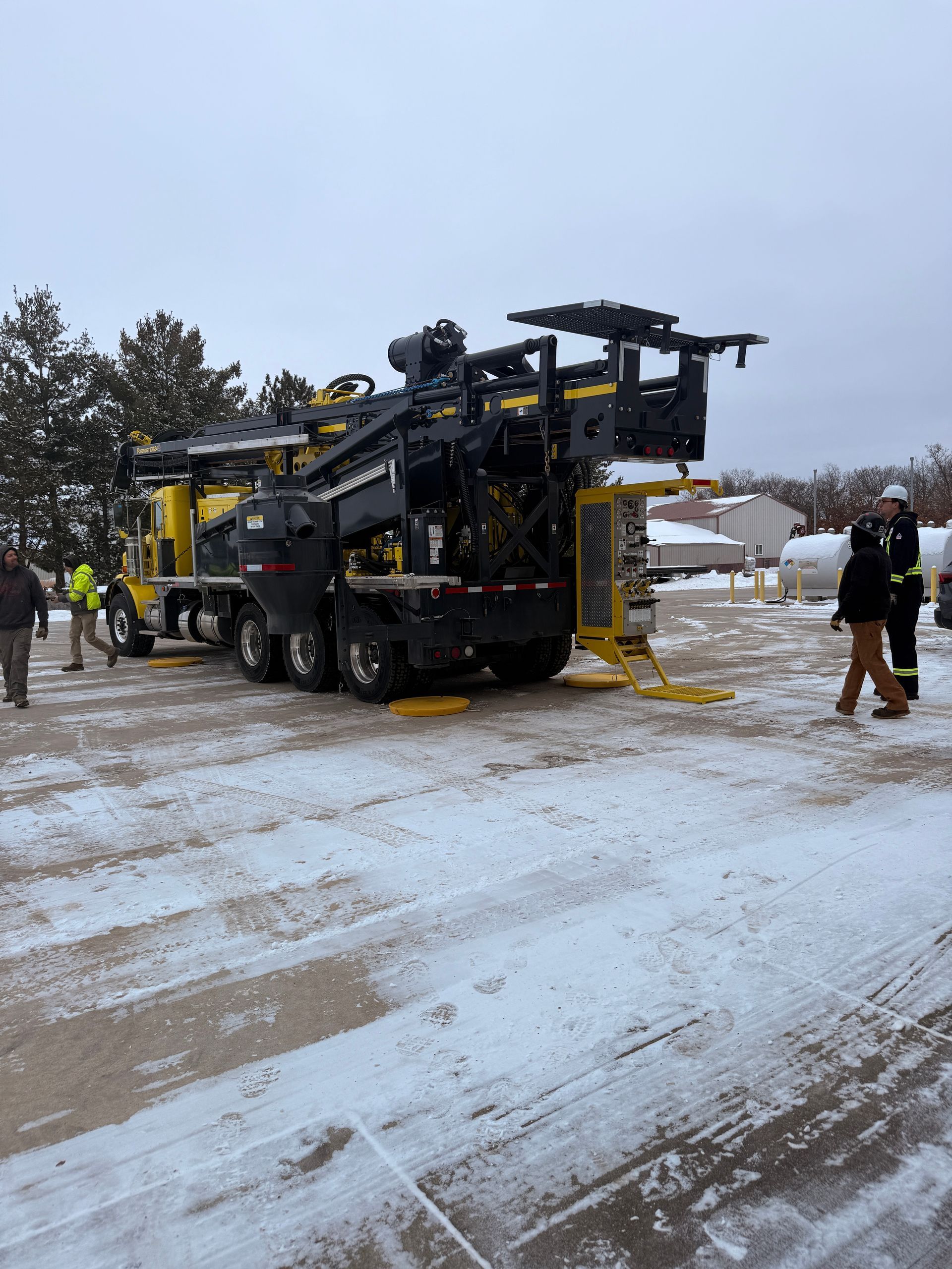 Yellow-and-black industrial machine on a snowy lot with a few people standing nearby