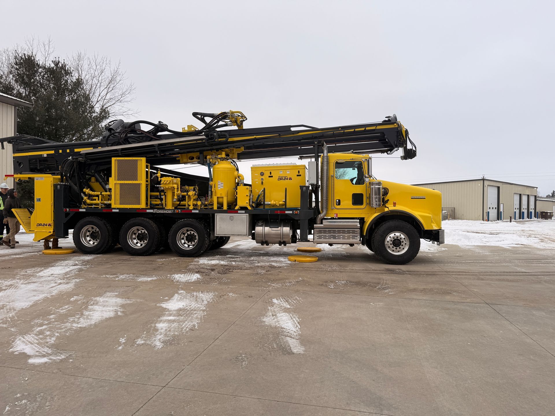 Yellow truck-mounted drilling rig parked on a snowy lot near industrial buildings