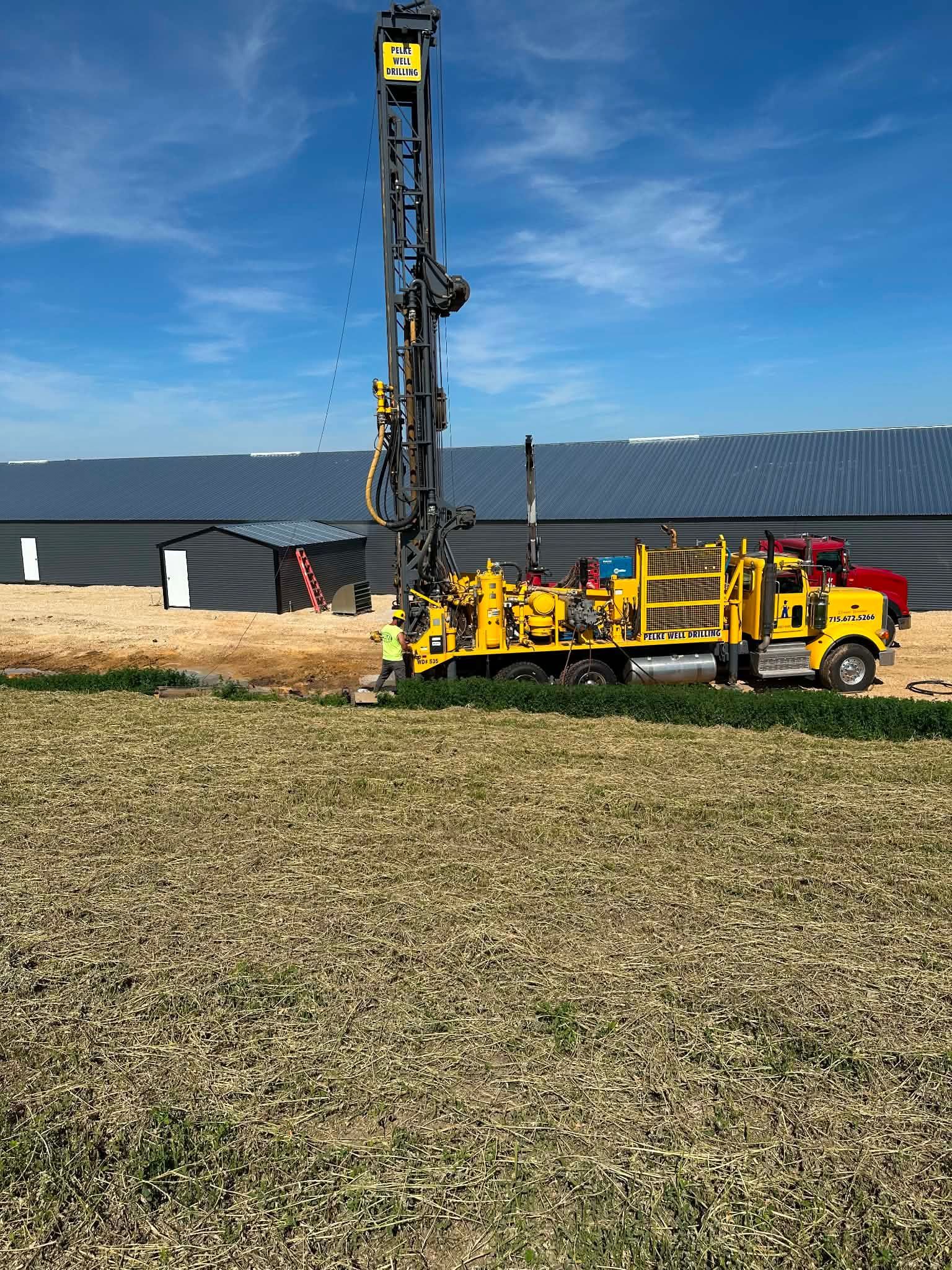 Yellow drilling rig truck in a grassy field near a dark building under a clear blue sky