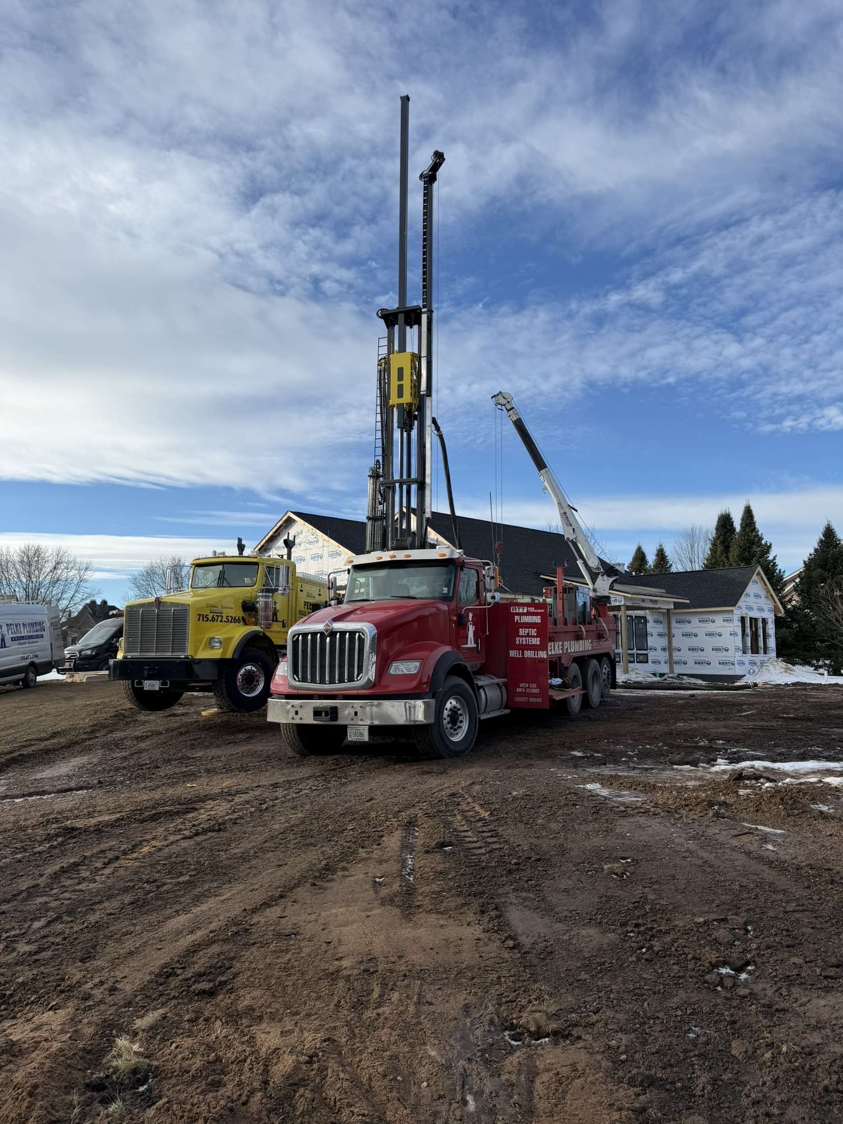 Drilling rig beside red and yellow construction trucks on a muddy site under a cloudy sky