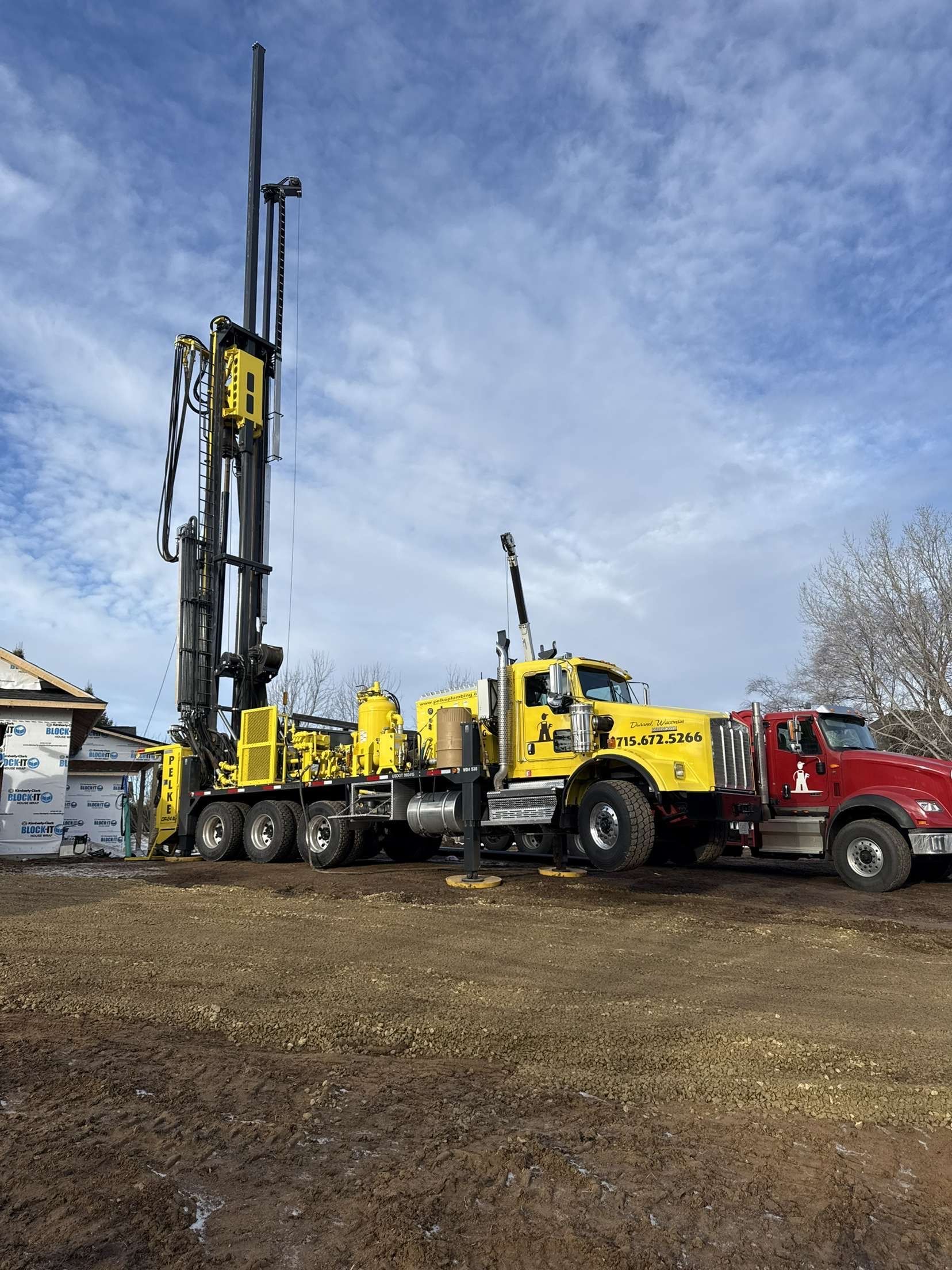 Yellow drilling rig truck beside a red pickup on a muddy construction site under a cloudy sky