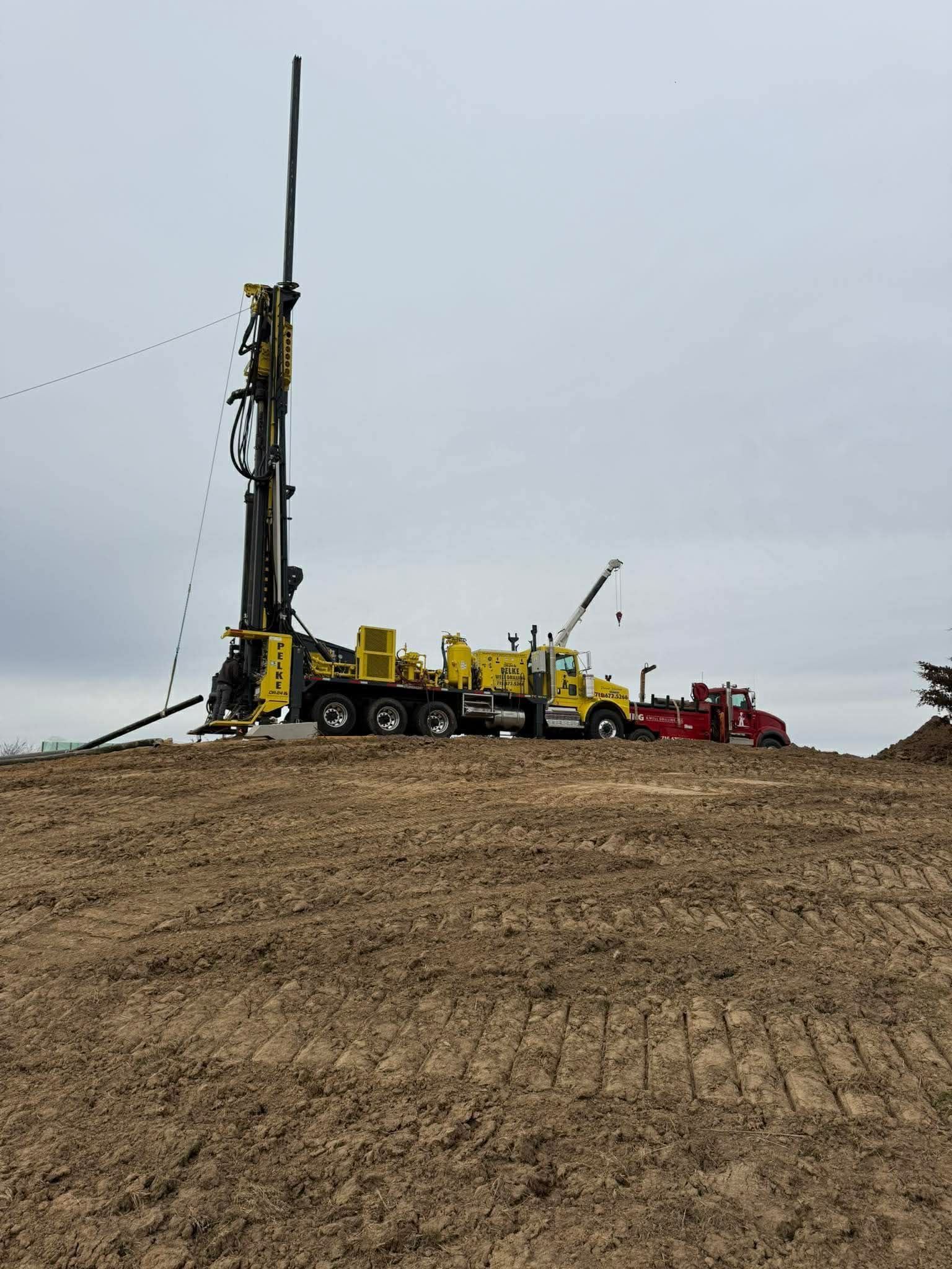 Yellow drilling rig and trucks on a dirt hill under an overcast sky.