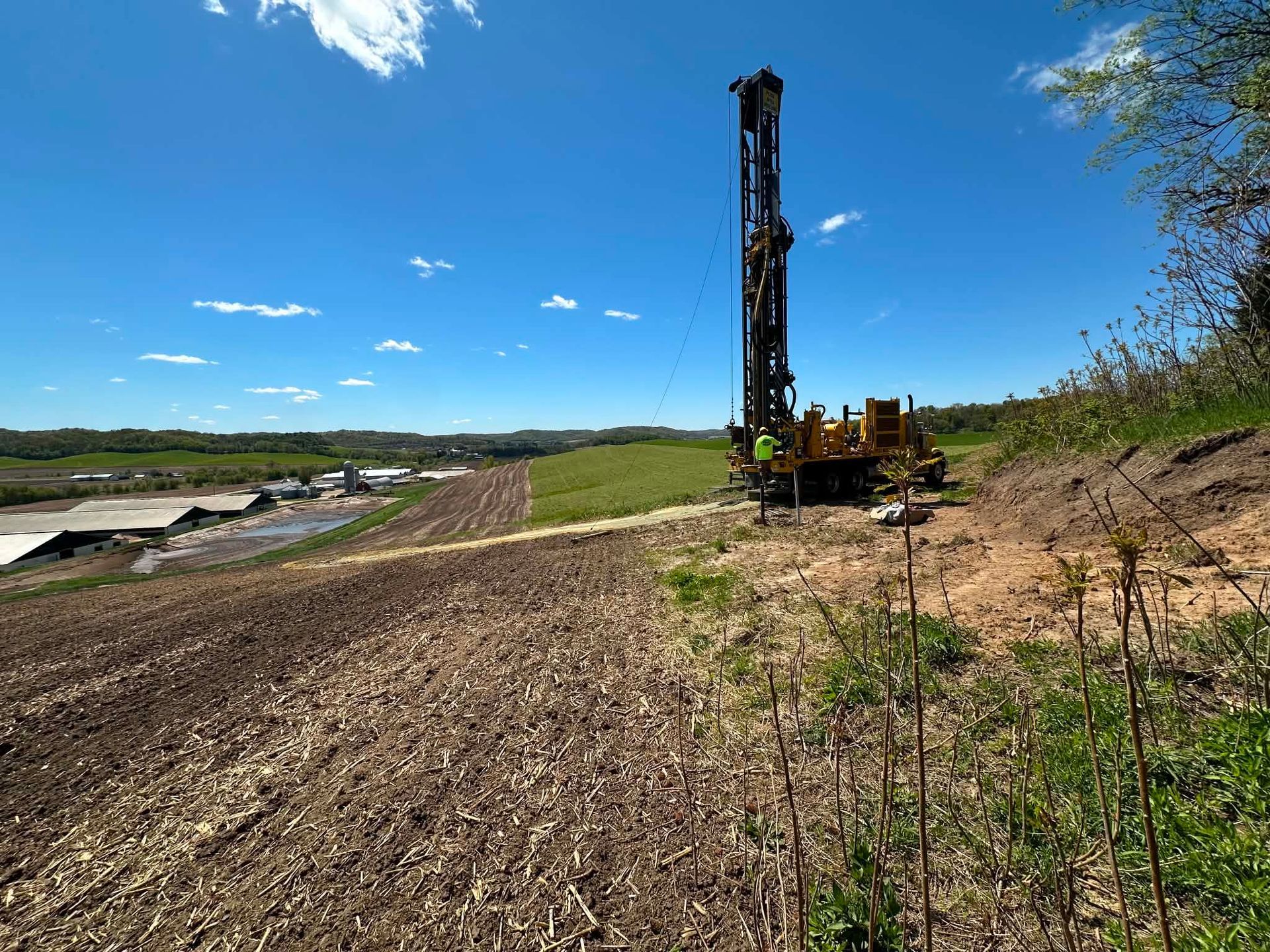 Drilling rig on a dirt road beside a grassy field under a blue sky