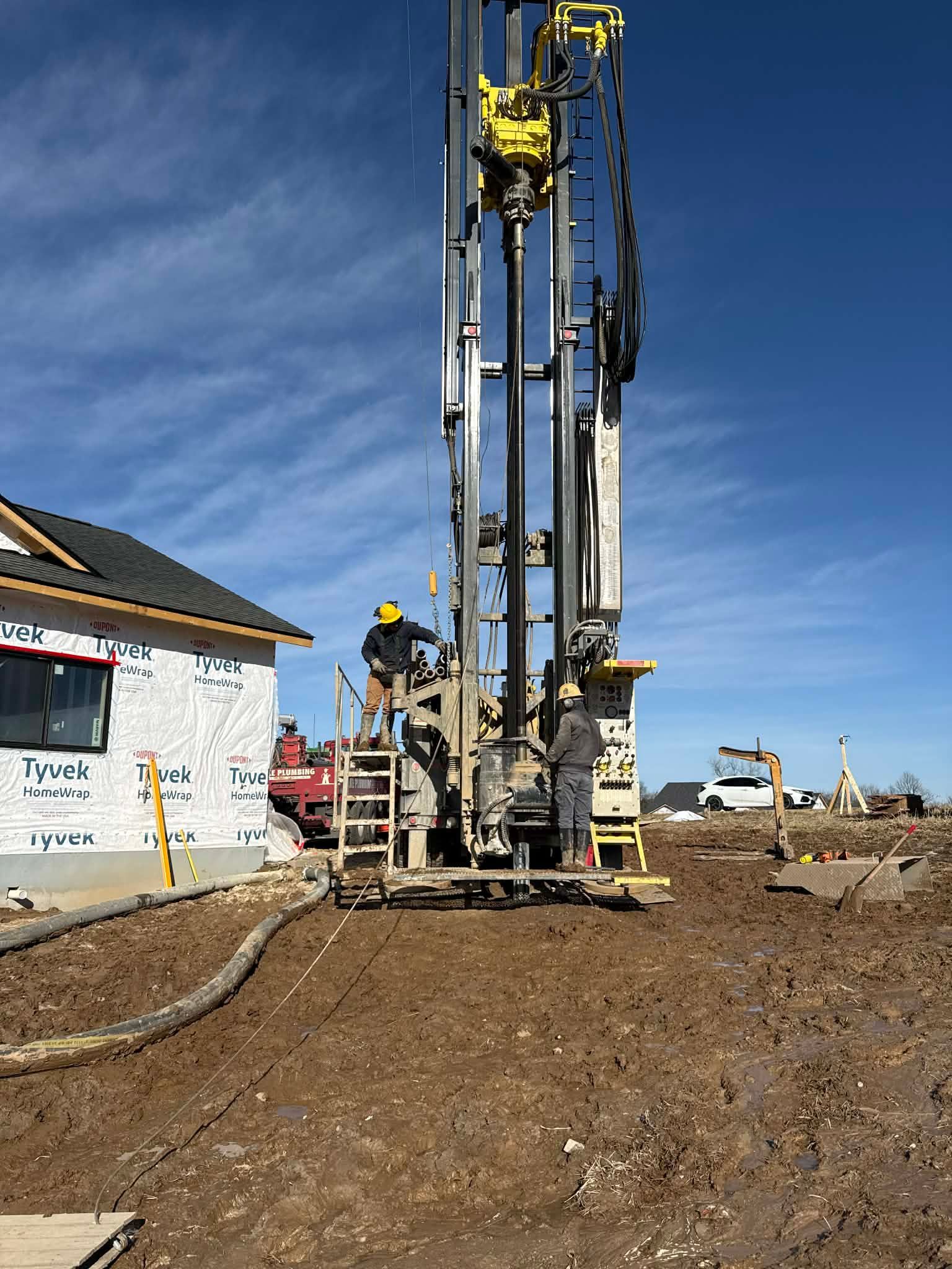 Drilling rig on a construction site beside a house, with workers and muddy ground under a blue sky