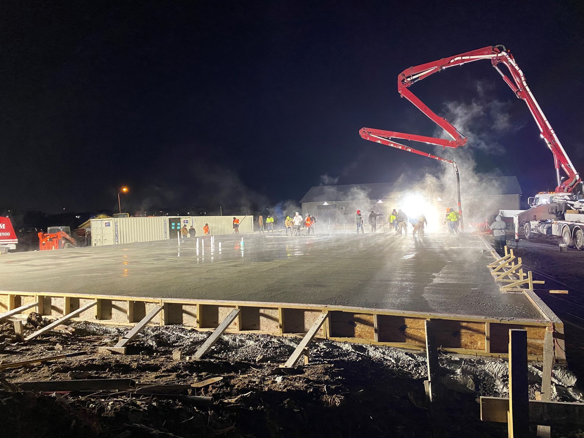 Night construction site: workers pouring concrete, illuminated by lights; large concrete pump.