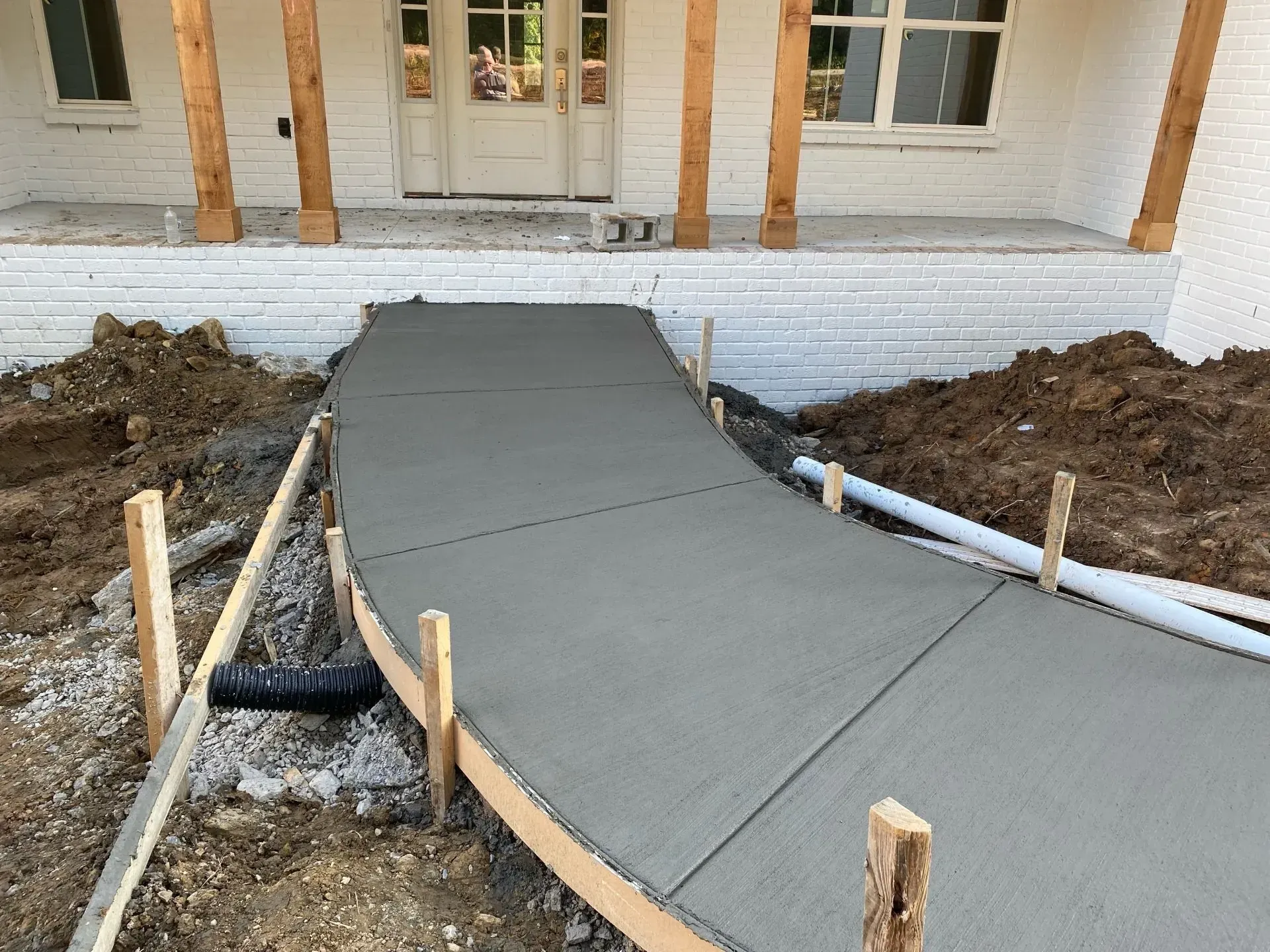 New concrete walkway being poured, leading to a white brick house entrance.