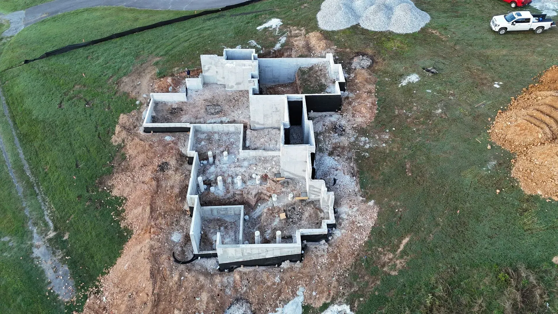 Aerial view of a concrete foundation under construction on a grassy lot, surrounded by dirt piles and a truck.