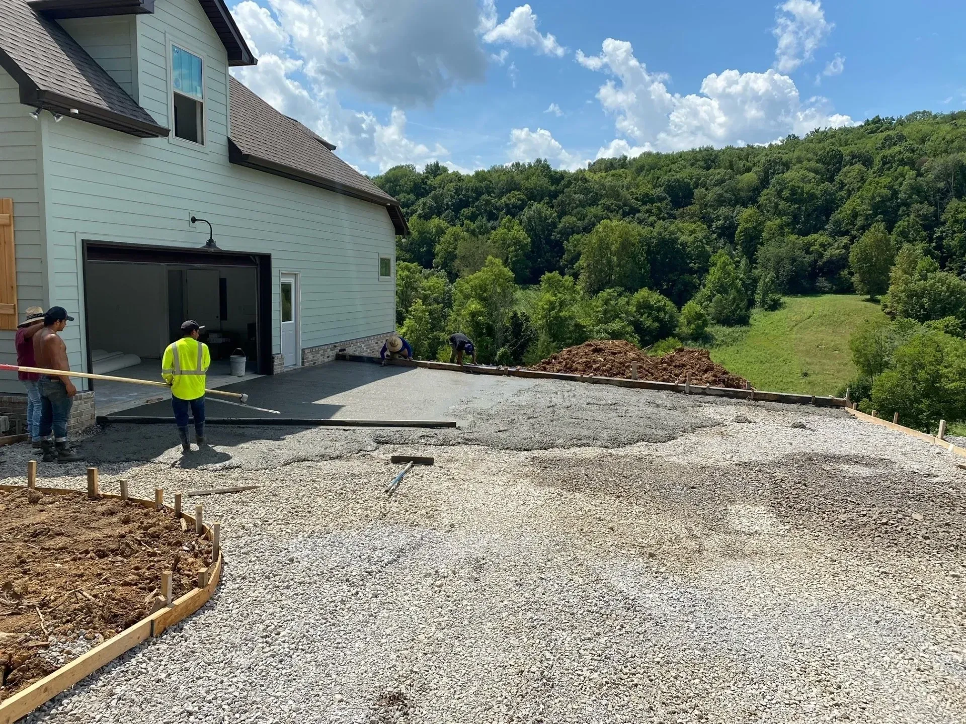 Concrete driveway being poured in front of a house under construction, with workers and a wooded hillside.