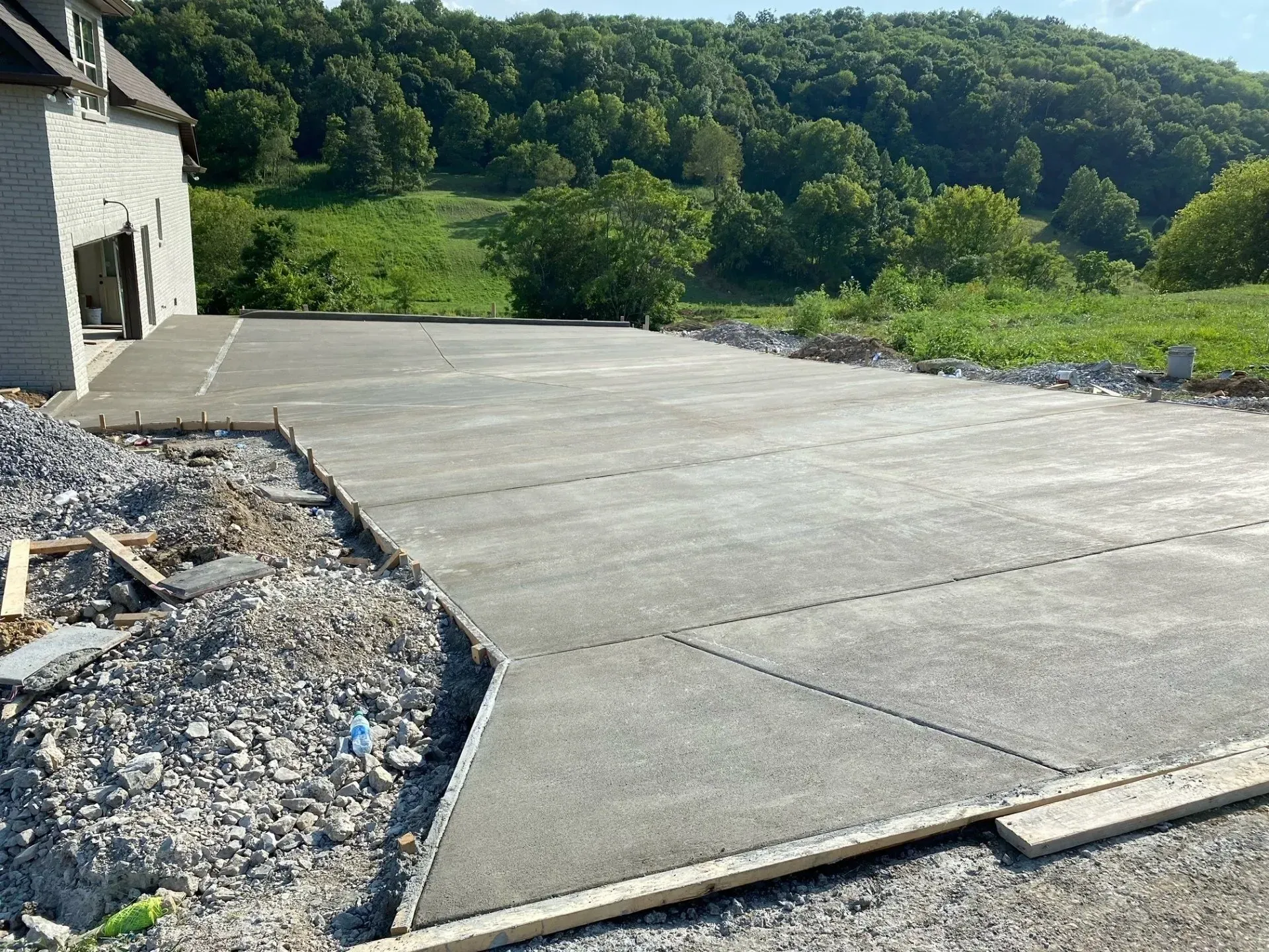 Newly poured concrete patio next to a light-colored house, with a gravel edge and a tree-filled hill in the background.