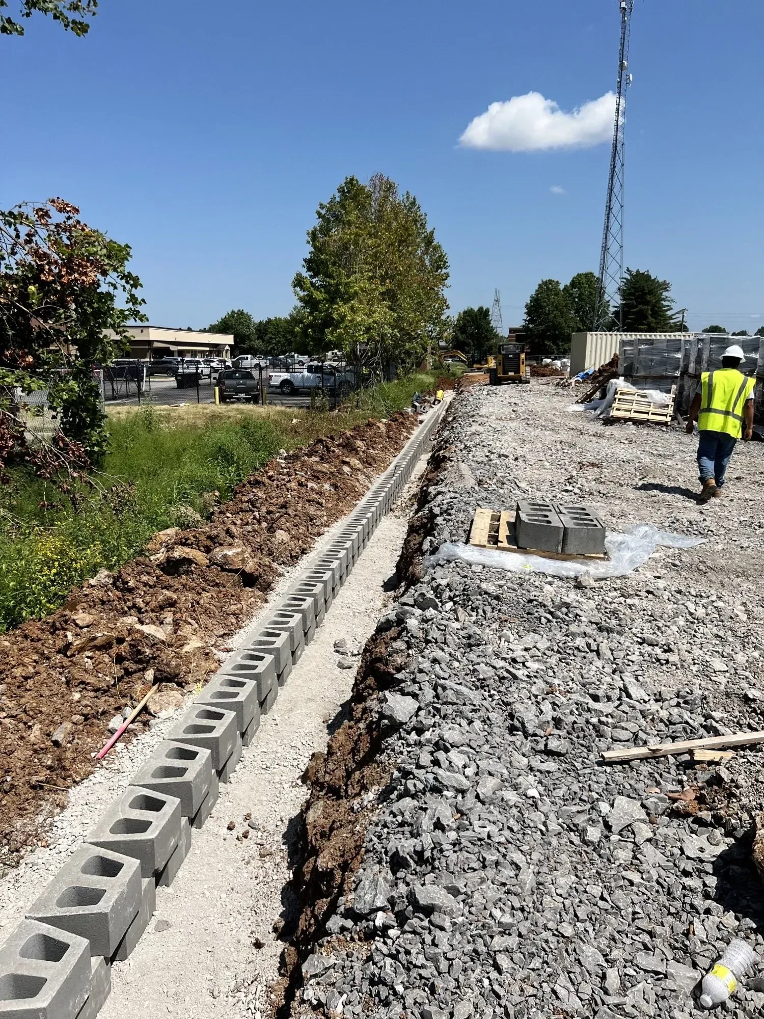 Construction site: blocks being laid for a wall. A worker in a vest walks by.
