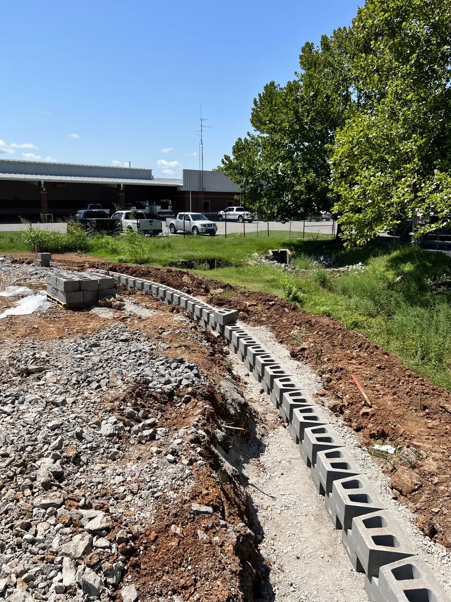 Construction site: cinder block retaining wall being built near a road and trees on a sunny day.