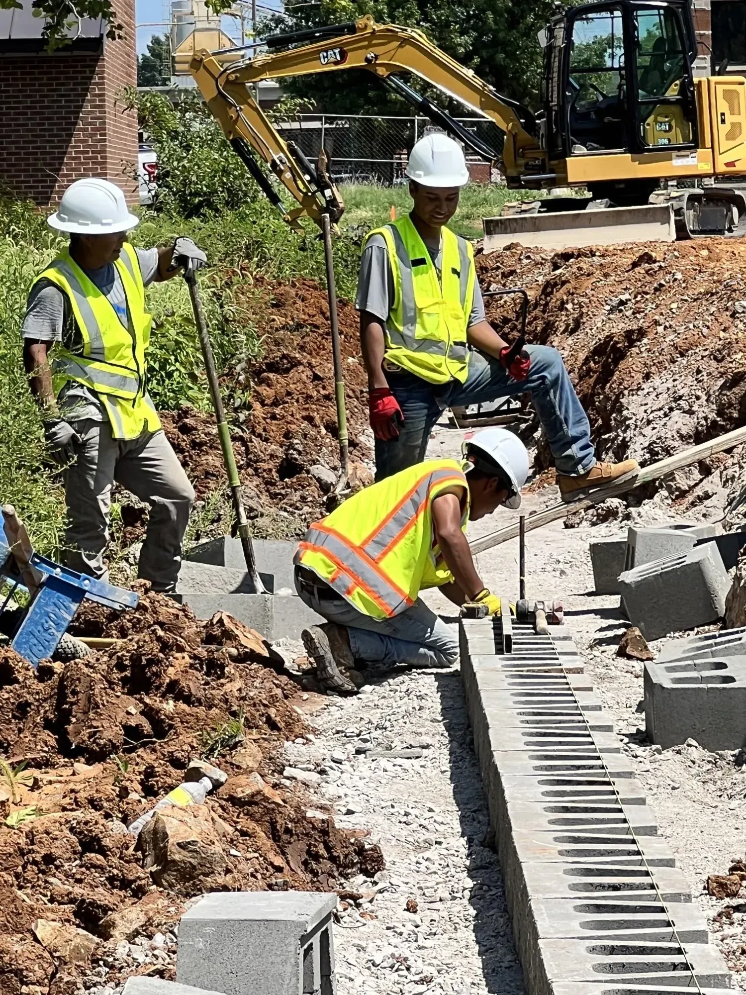 Construction workers install pavers; yellow vests, hard hats, digging, kneeling.