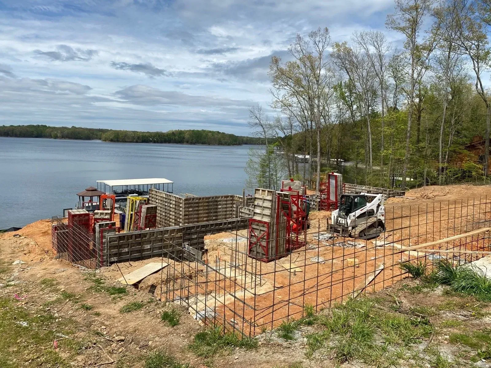 Construction site by a lake; forms, rebar, and equipment are set for a concrete foundation.