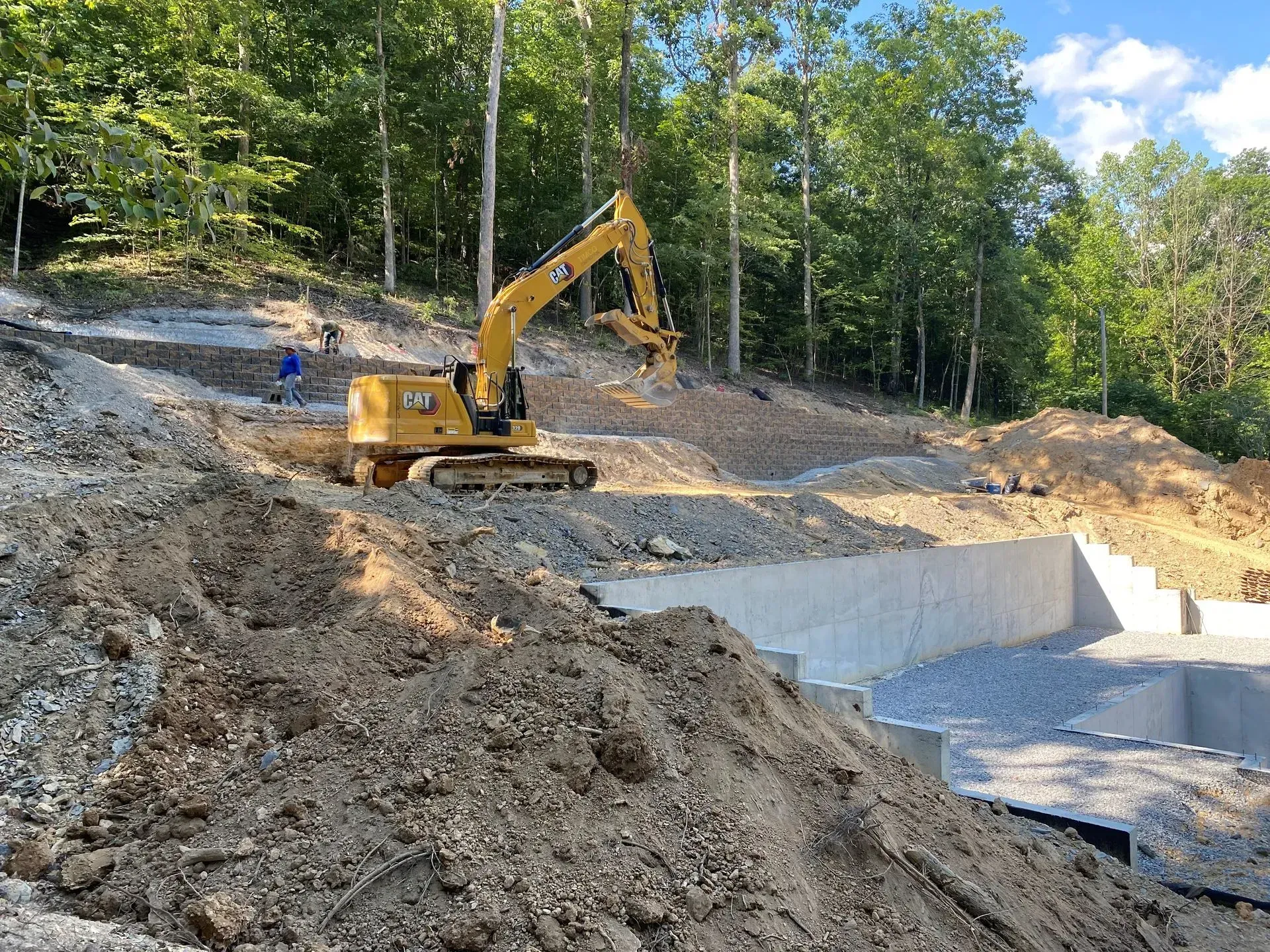 Yellow excavator grading a hillside construction site near a concrete retaining wall. Forest background.