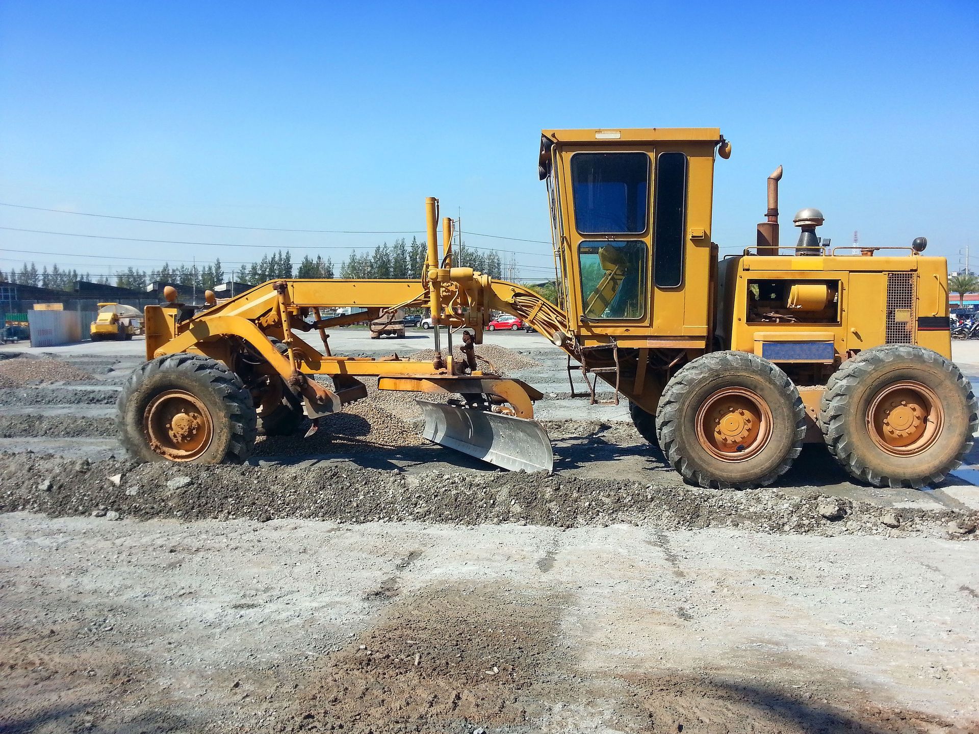 Yellow road grader smoothing gravel on a construction site under a blue sky.