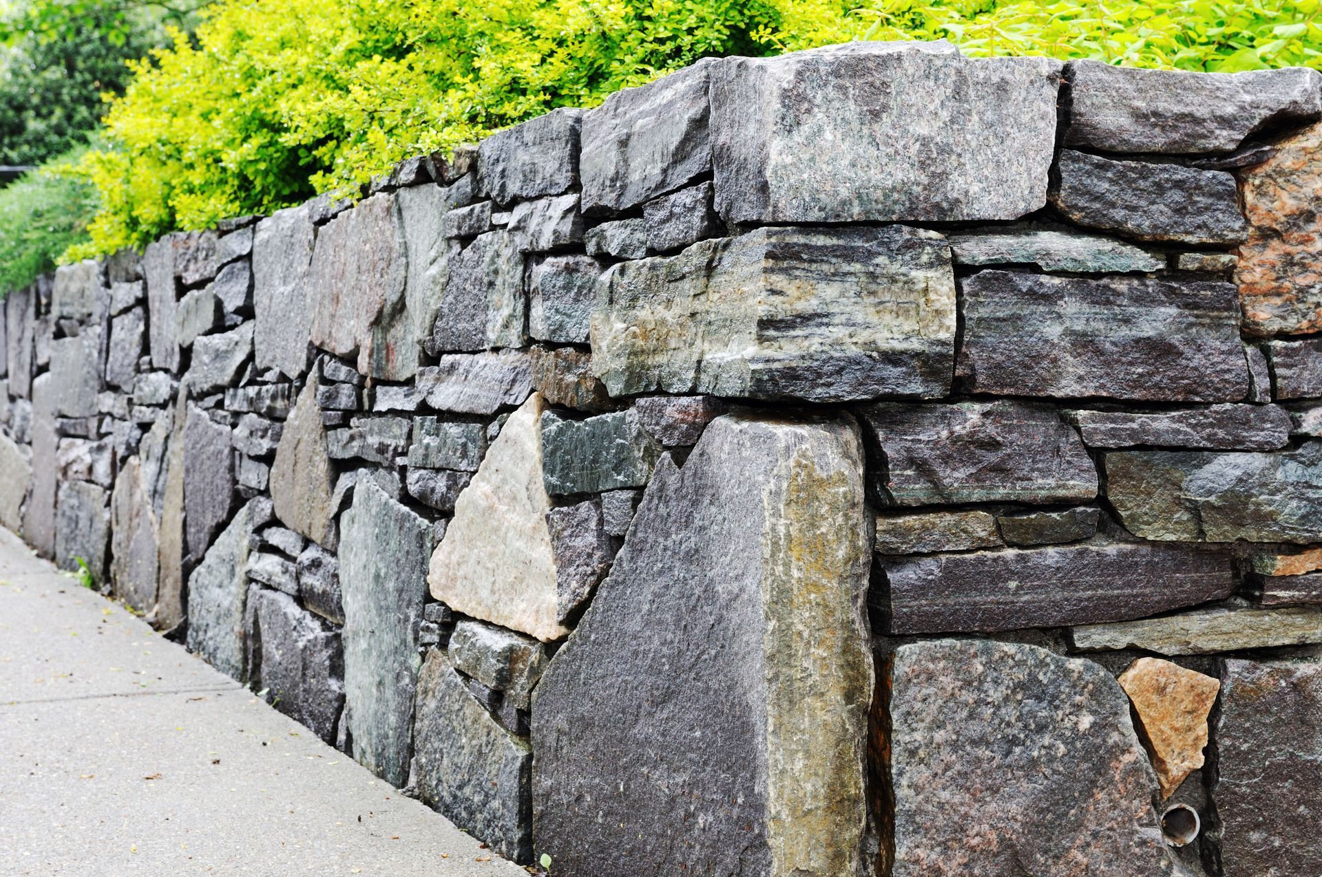 Stone wall with mixed gray, brown, and beige rocks, bordering a walkway with green foliage on top.