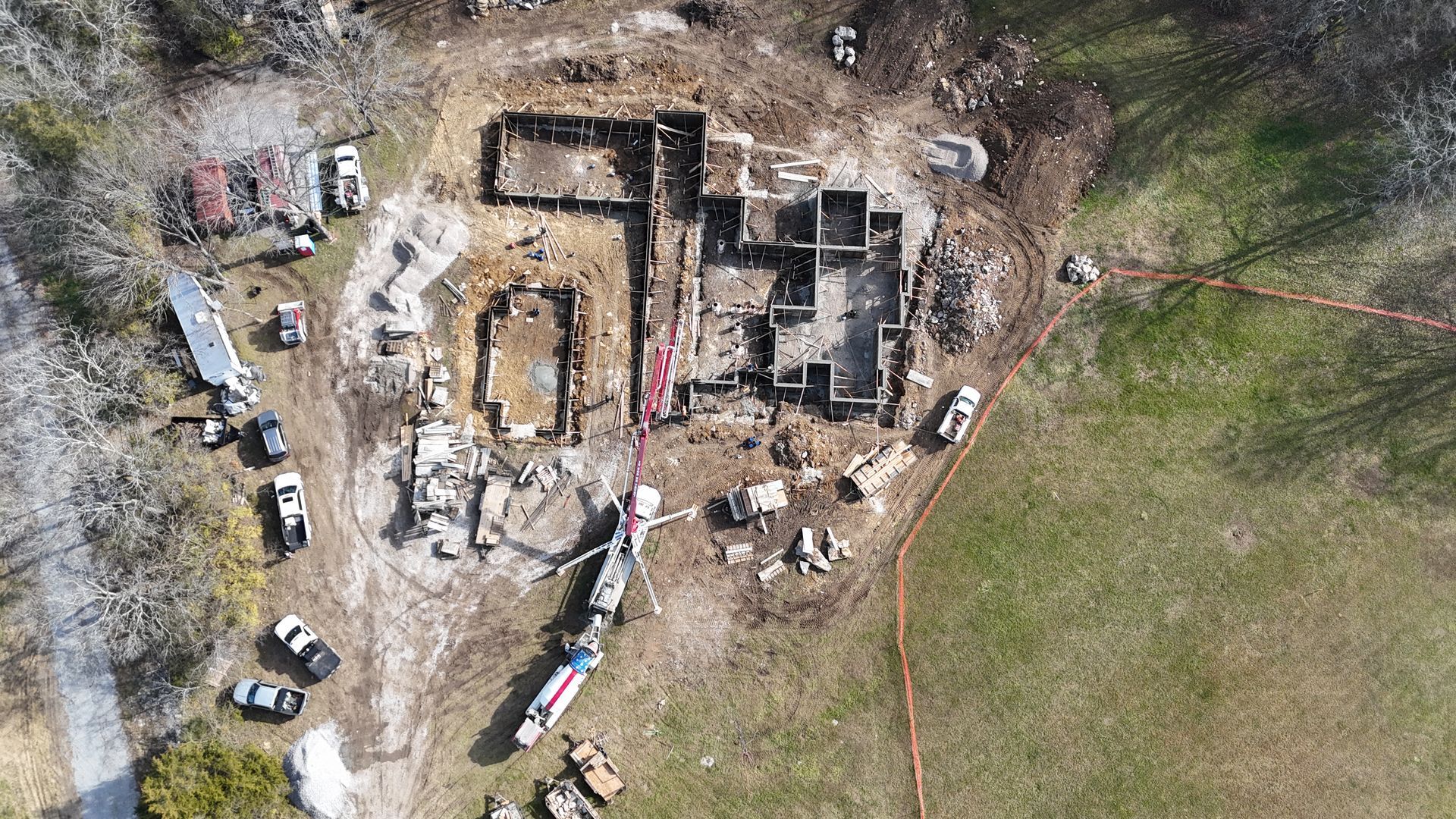 Aerial view of a partially demolished building site with foundation remains, surrounded by construction vehicles, debris, and green grass.